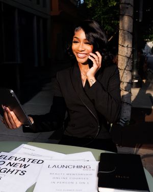 A person in a black jacket smiles while sitting at a desk with documents in a dimly lit setting.