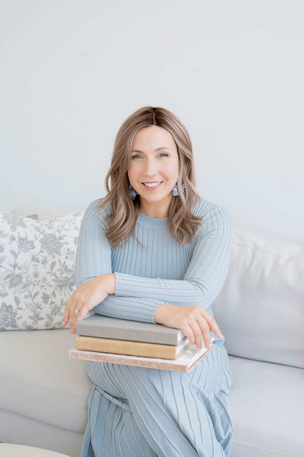 Woman in blue sweater poses with books on light gray couch in bright interior.