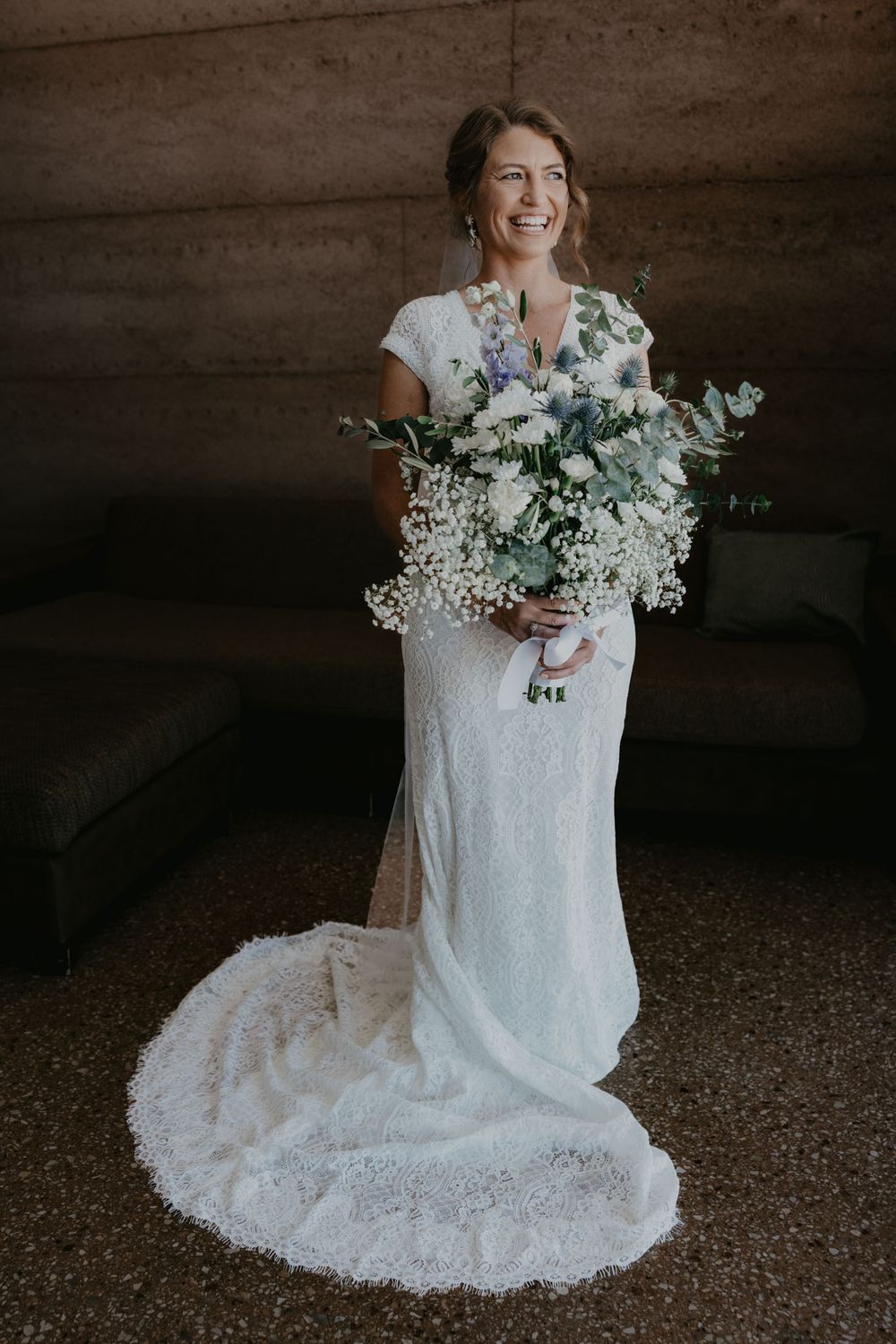 A bride in a white lace wedding dress holds a delicate bouquet of baby's breath flowers against a dark background.