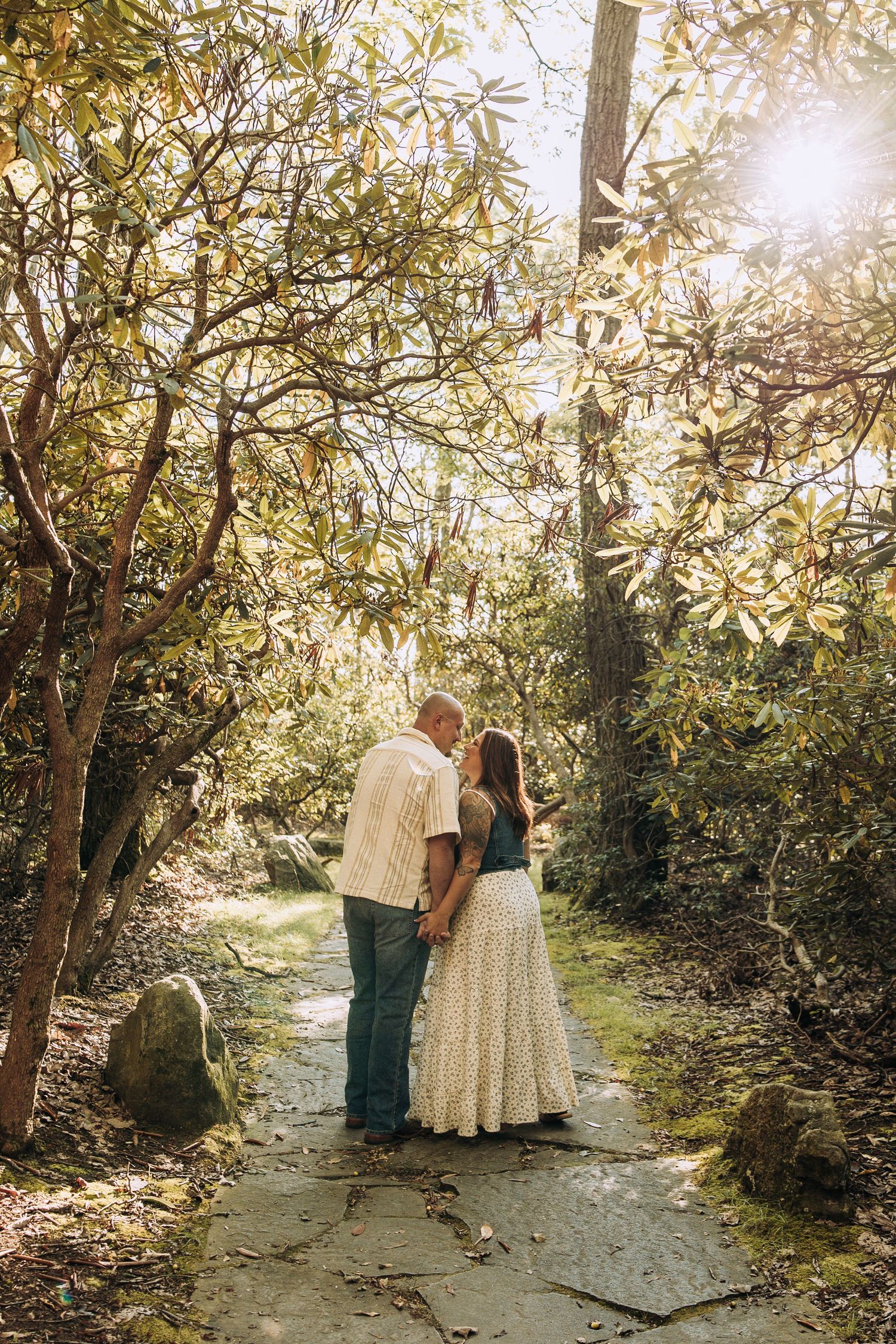 Couple sharing romantic moment on stone path surrounded by lush green trees in sunlit garden.