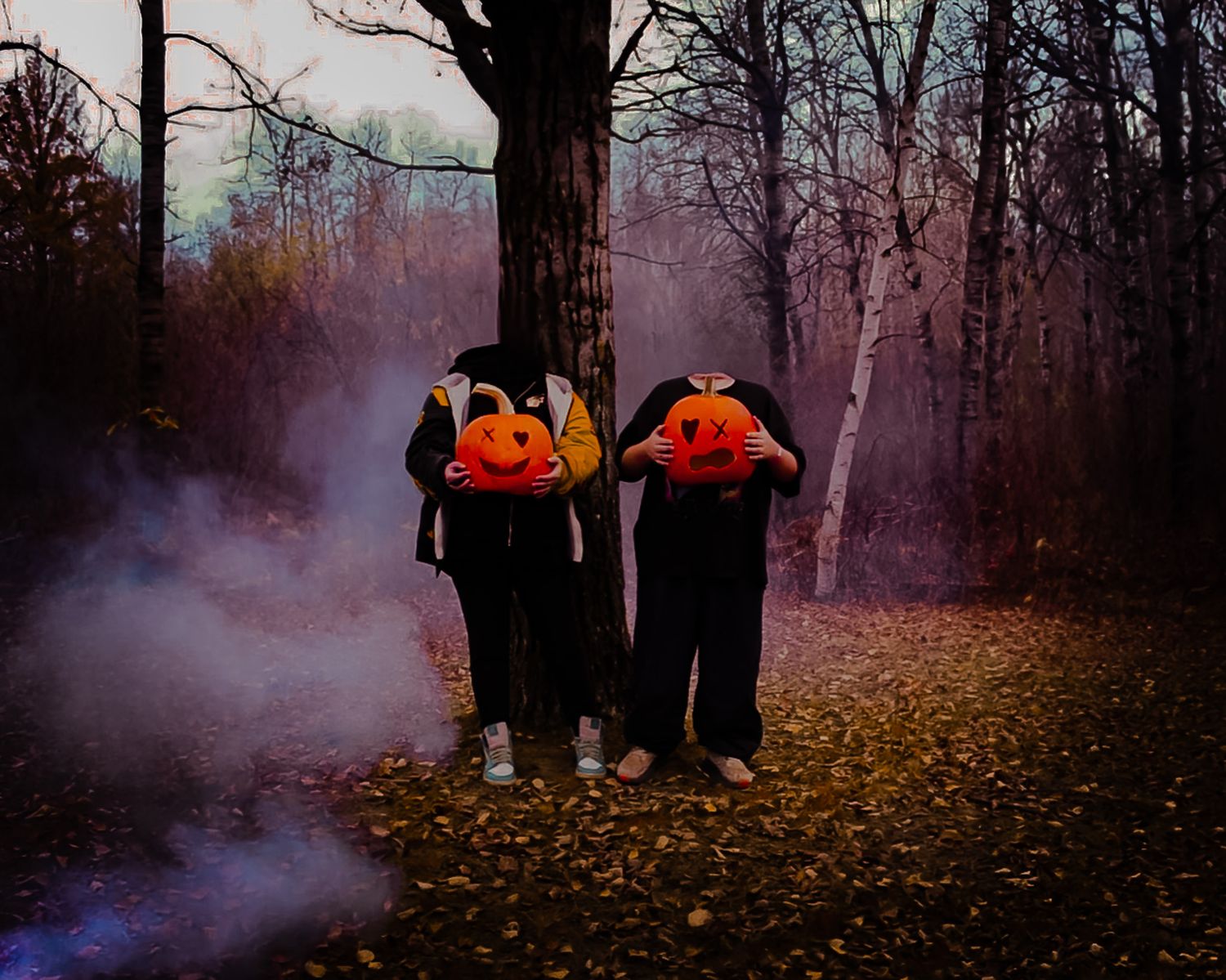 Two figures in black clothing carry orange pumpkins while walking through a misty autumn forest with fallen leaves.