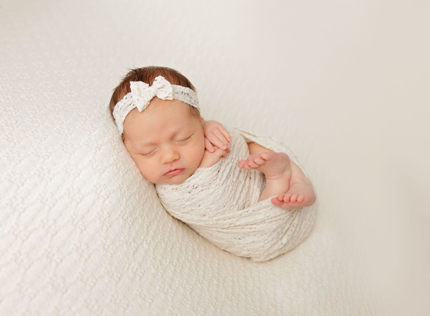A sleeping newborn baby wrapped in soft white fabric lies peacefully on a white textured background.