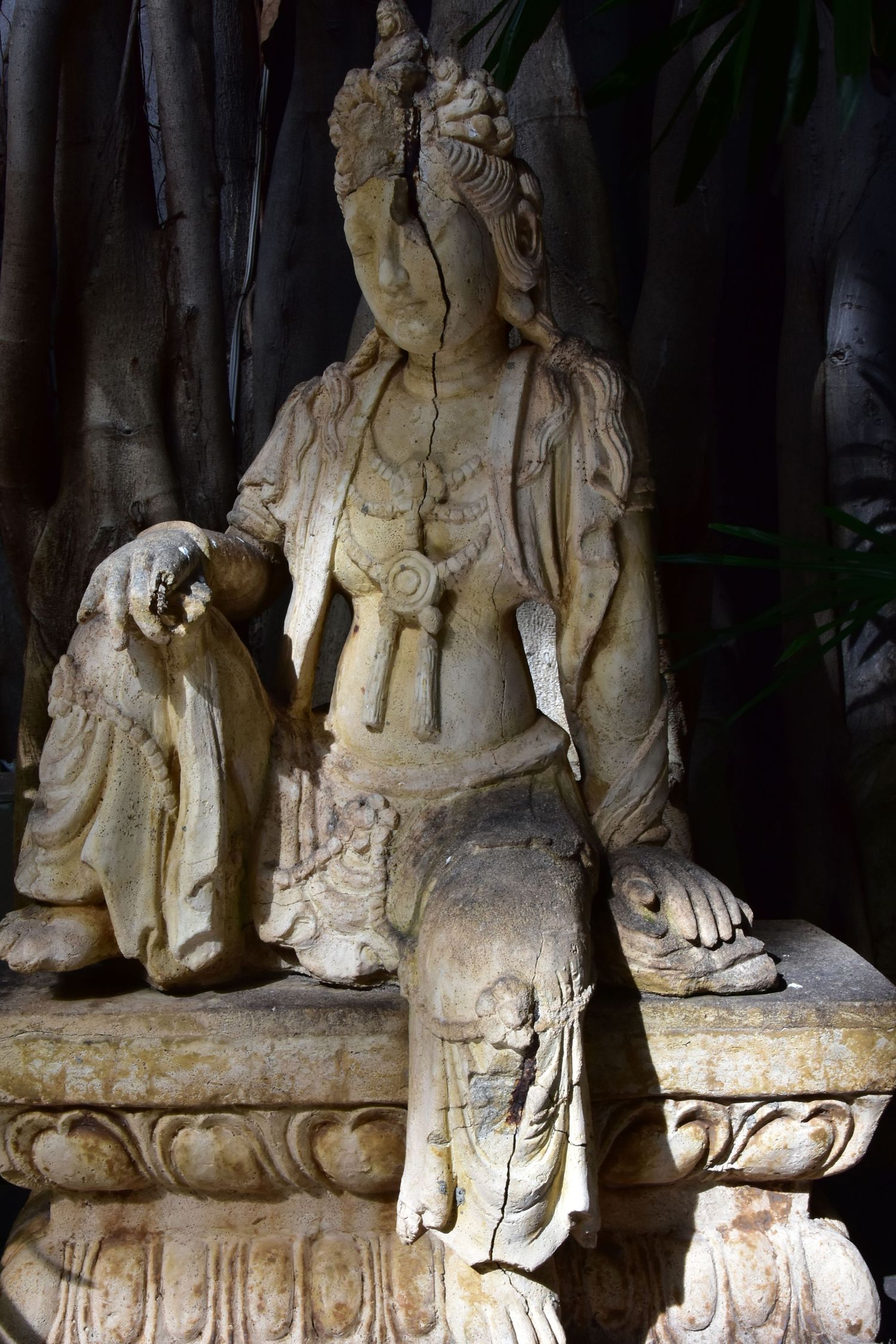 An ancient eastern style statue sits on a bench under a tree.  Colour version.