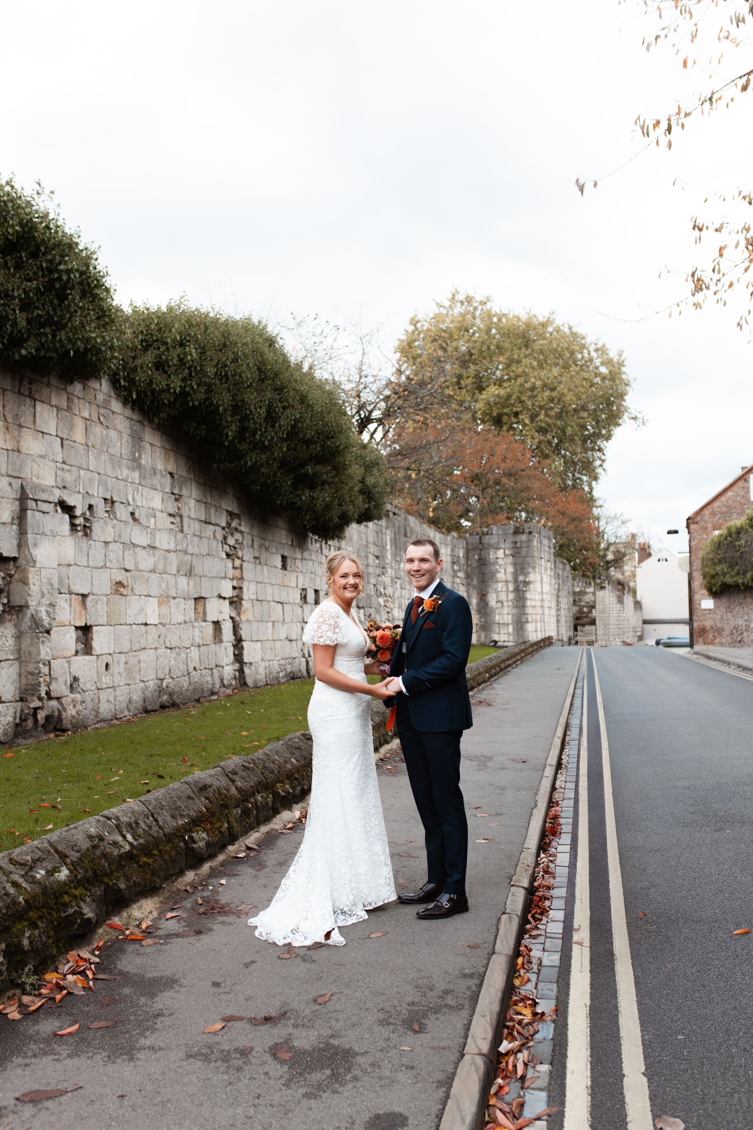 A couple in wedding attire stands together on a quiet street with stone walls and autumn leaves scattered along the curb.