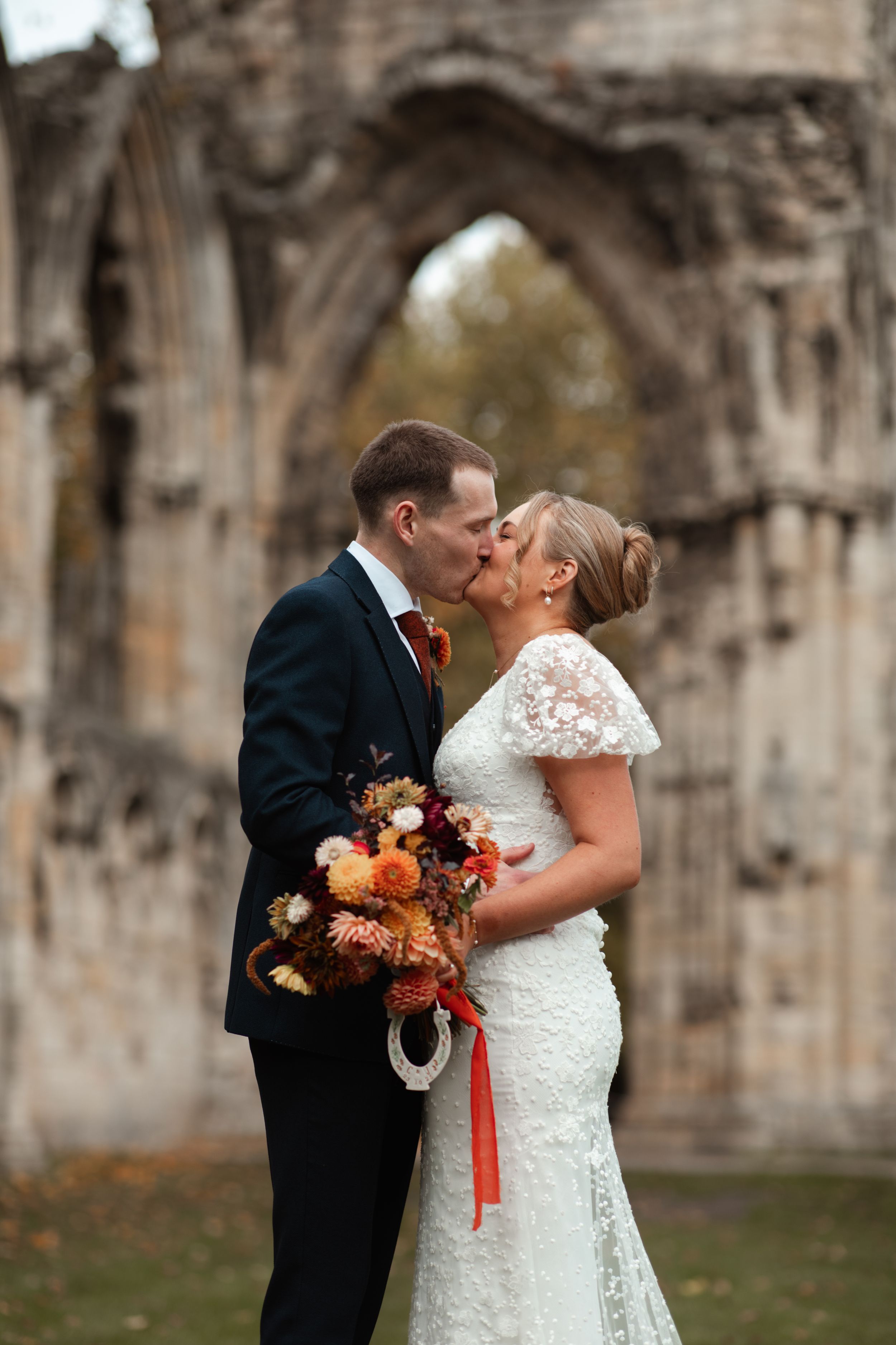 A romantic wedding couple embraces in front of a rustic stone wall on their autumn wedding day.