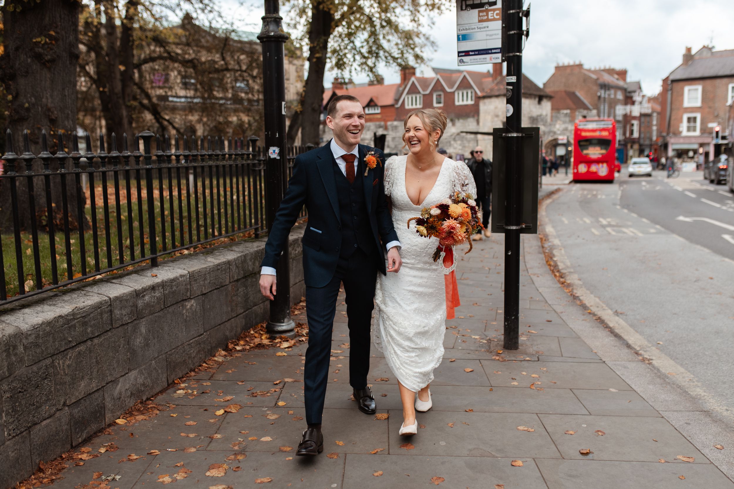 Wedding party walks down a city sidewalk during autumn with fallen leaves scattered on the ground.