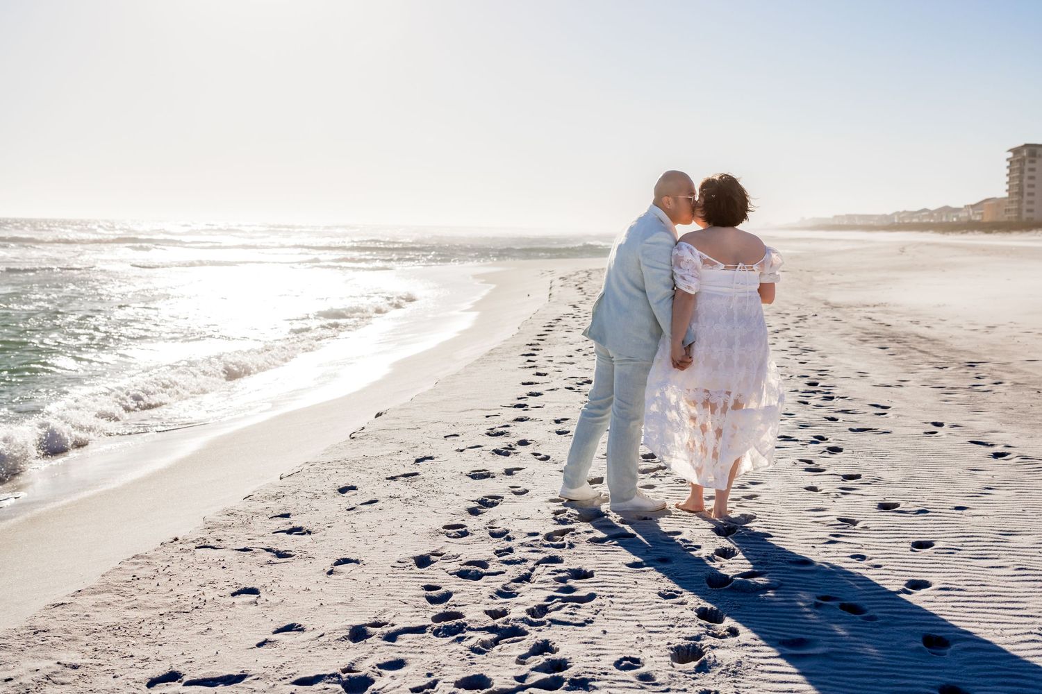 A couple walks along a sandy beach at sunset leaving footprints in their wake as waves crash nearby.