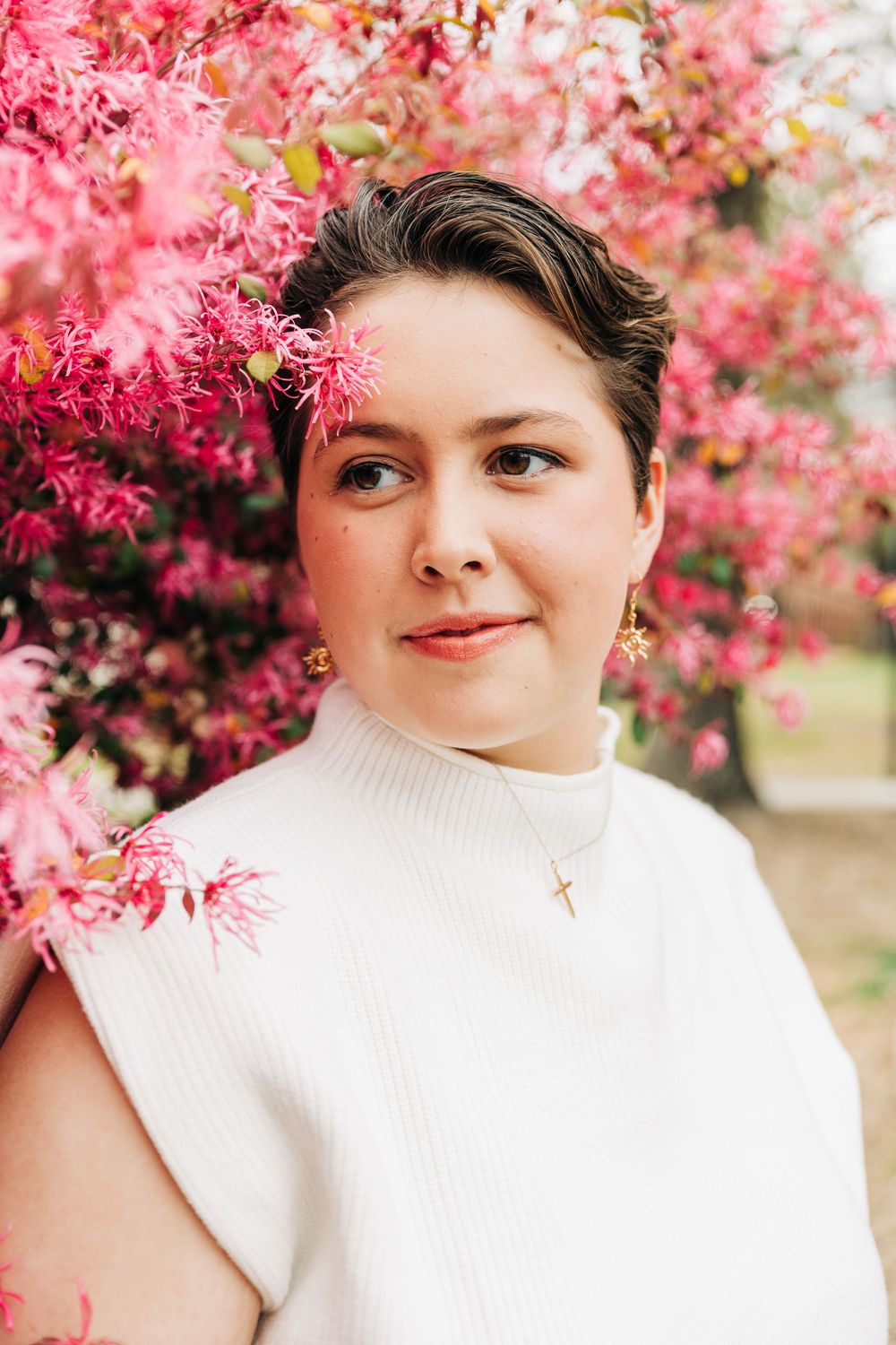 Person in white turtleneck sweater poses against vibrant pink spring blossoms in outdoor portrait setting in Matthews, NC.