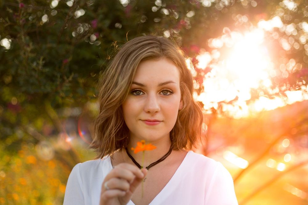 A female in a white top poses outdoors during golden hour sunset with glowing bokeh and yellow wildflowers in background of Camp North End