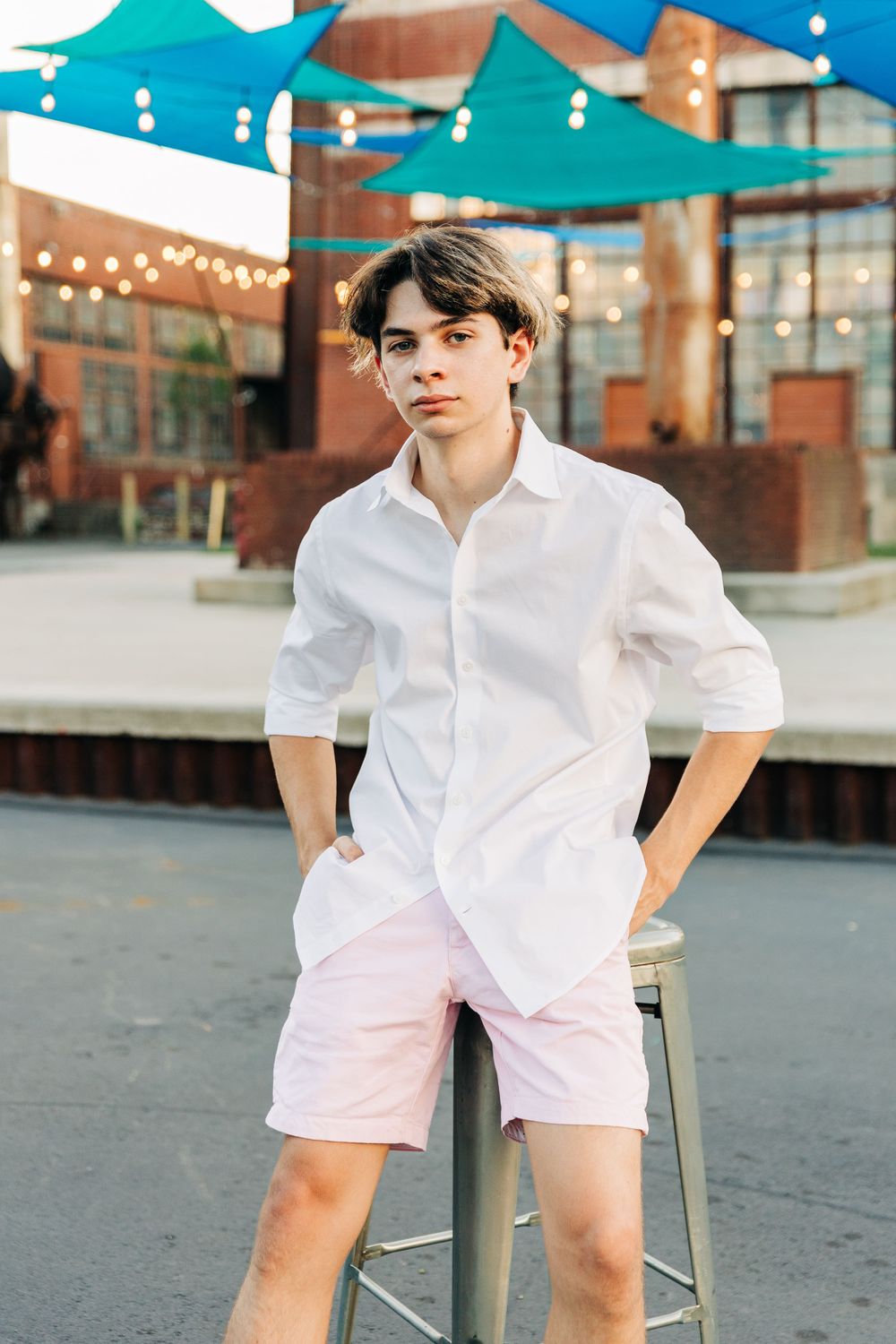 Young male in white button-down shirt and pink shorts sitting casually on stool outdoors with teal awning and string lights in Charlotte.
