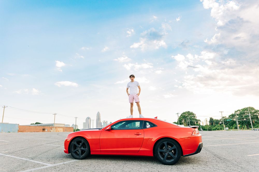 A bright red sports car with a male standing on top of it in the Camp North End parking lot with dramatic cloudy skies in the background.