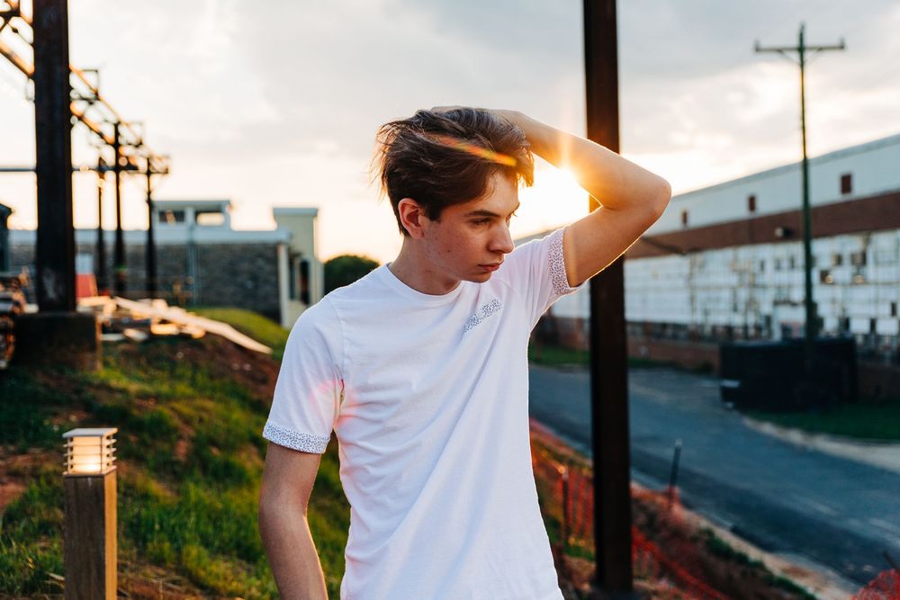 Person in white t-shirt stands against sunset sky with mountains and urban landscape in background.