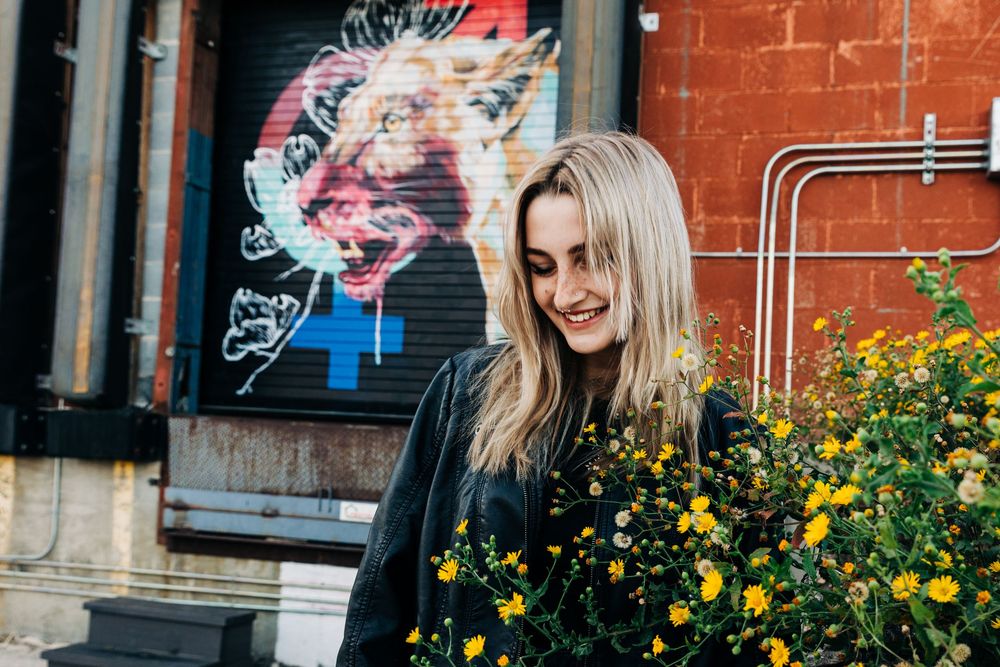 A girl with blonde hair smiles while standing next to yellow flowers outside a brick building for Charlotte senior photos.