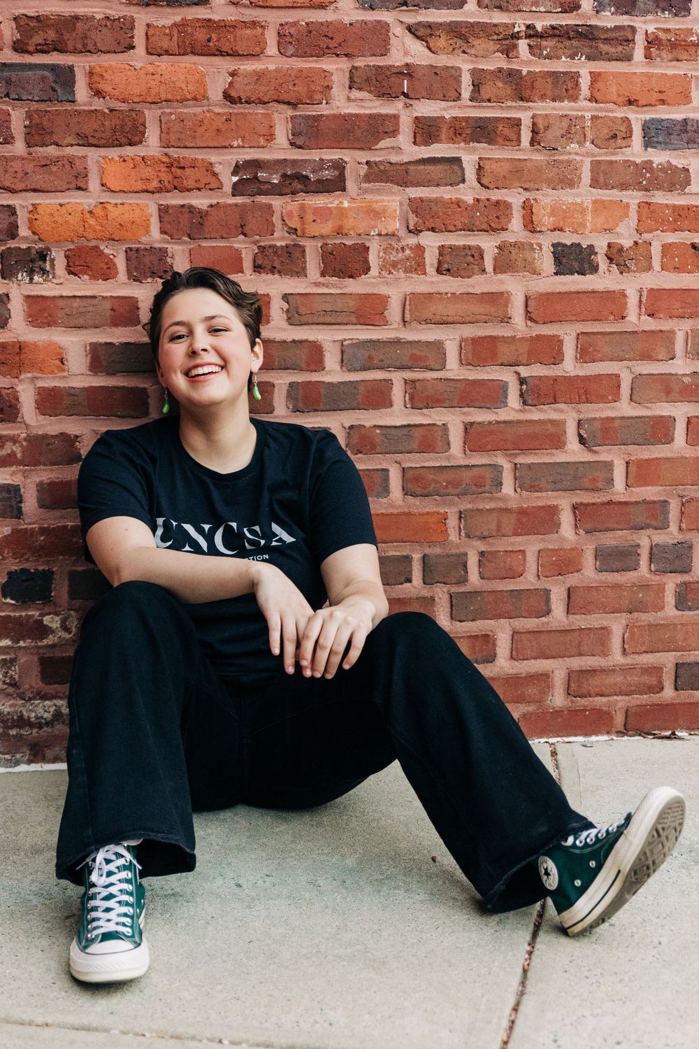 A person wearing black clothing and sneakers sits casually against a red brick wall for Charlotte senior Portraits.