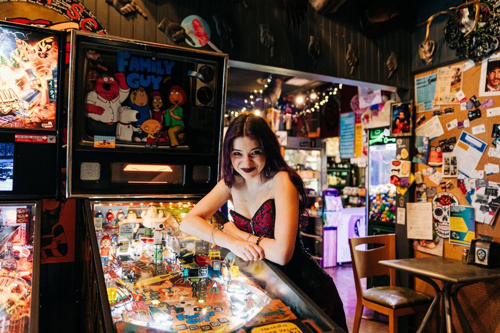 A girl leans over a game in a dimly lit space decorated with colorful photos and string lights on the walls for Monroe senior portraits.