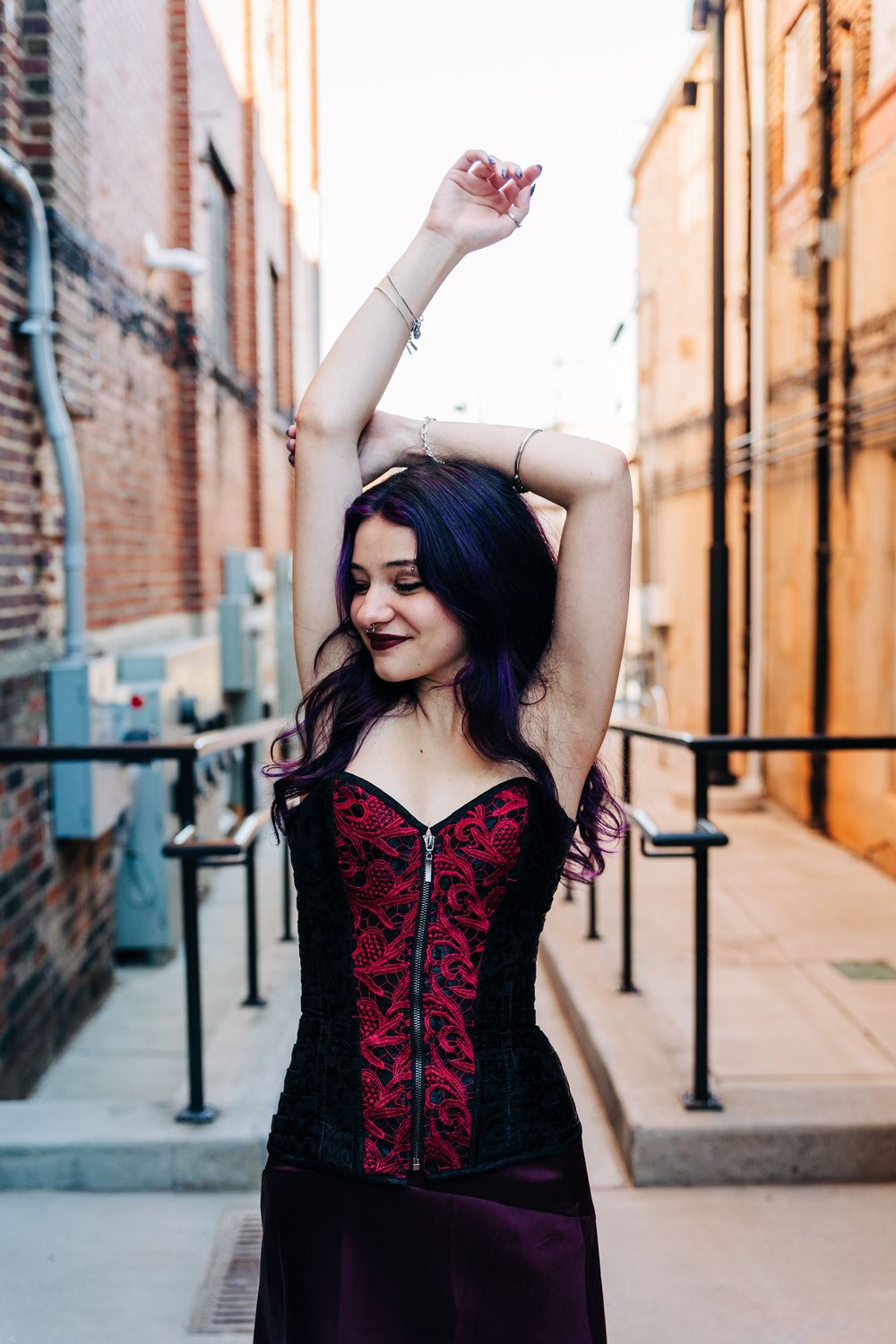 A woman in a burgundy corset dress poses confidently in an urban alleyway with brick walls and metal posts in Monroe, NC.