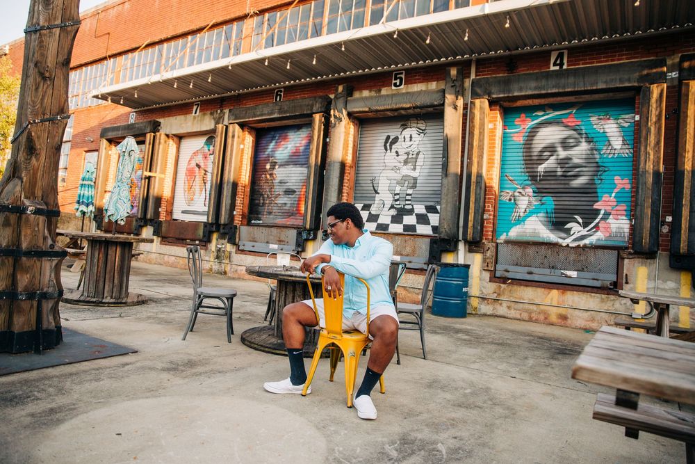 Someone sits on a chair outside an urban storefront decorated with colorful portrait murals and artwork for Charlotte senior portraits.