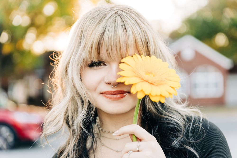 A bright yellow sunflower held up against a girls face outdoors with soft bokeh lighting in Monroe, NC.