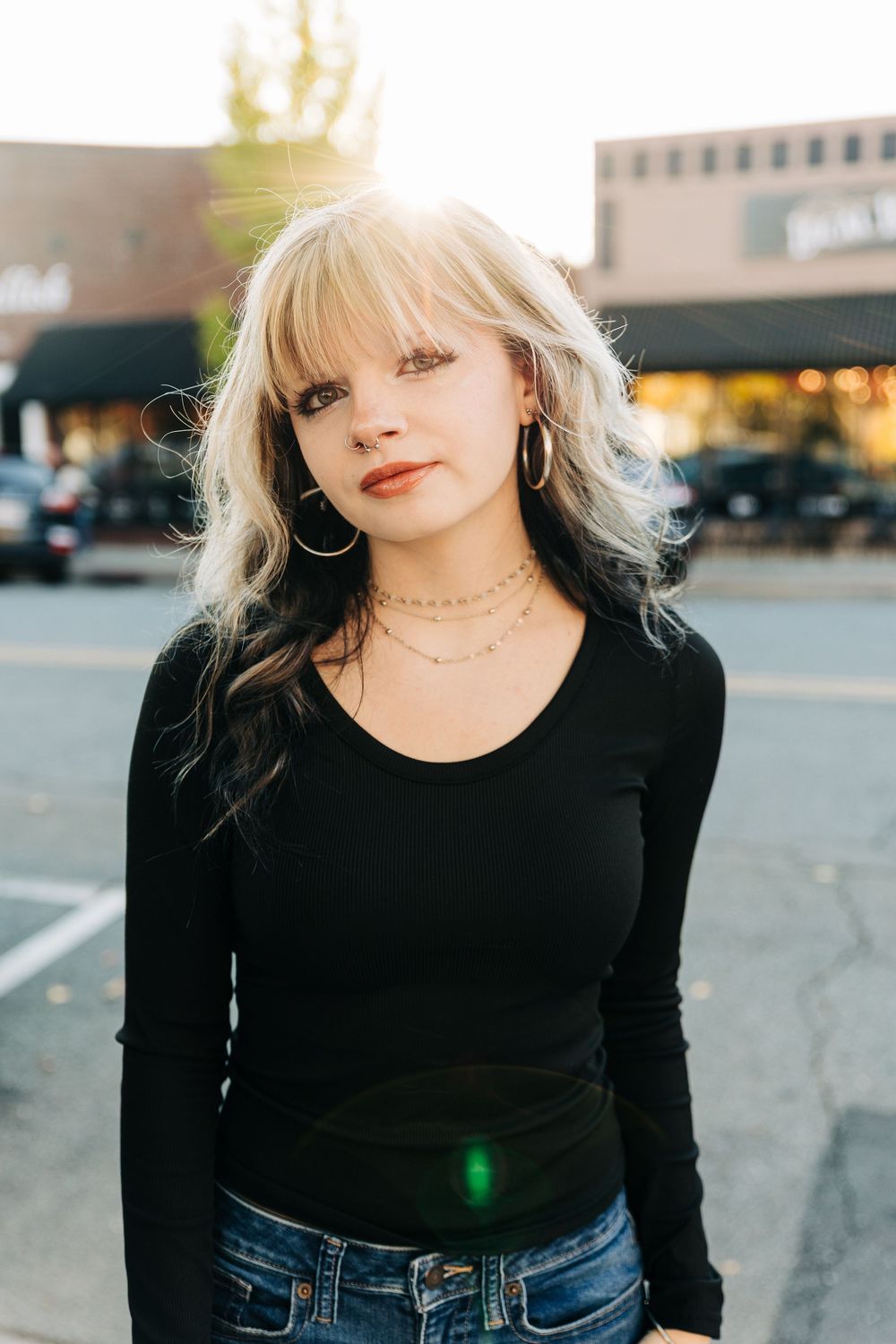 Person wearing black long sleeve top and blue jeans poses in parking lot during sunset.