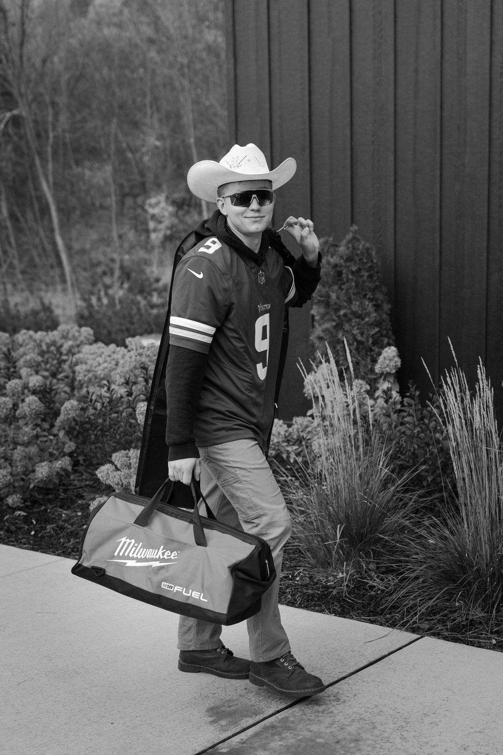 Person in cowboy hat and sunglasses walking with Milwaukee tool bags in black and white photo.