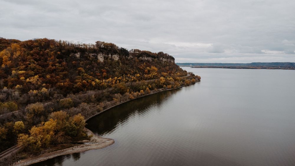 Aerial view of a rocky bluff with autumn foliage overlooking a calm lake under overcast skies.
