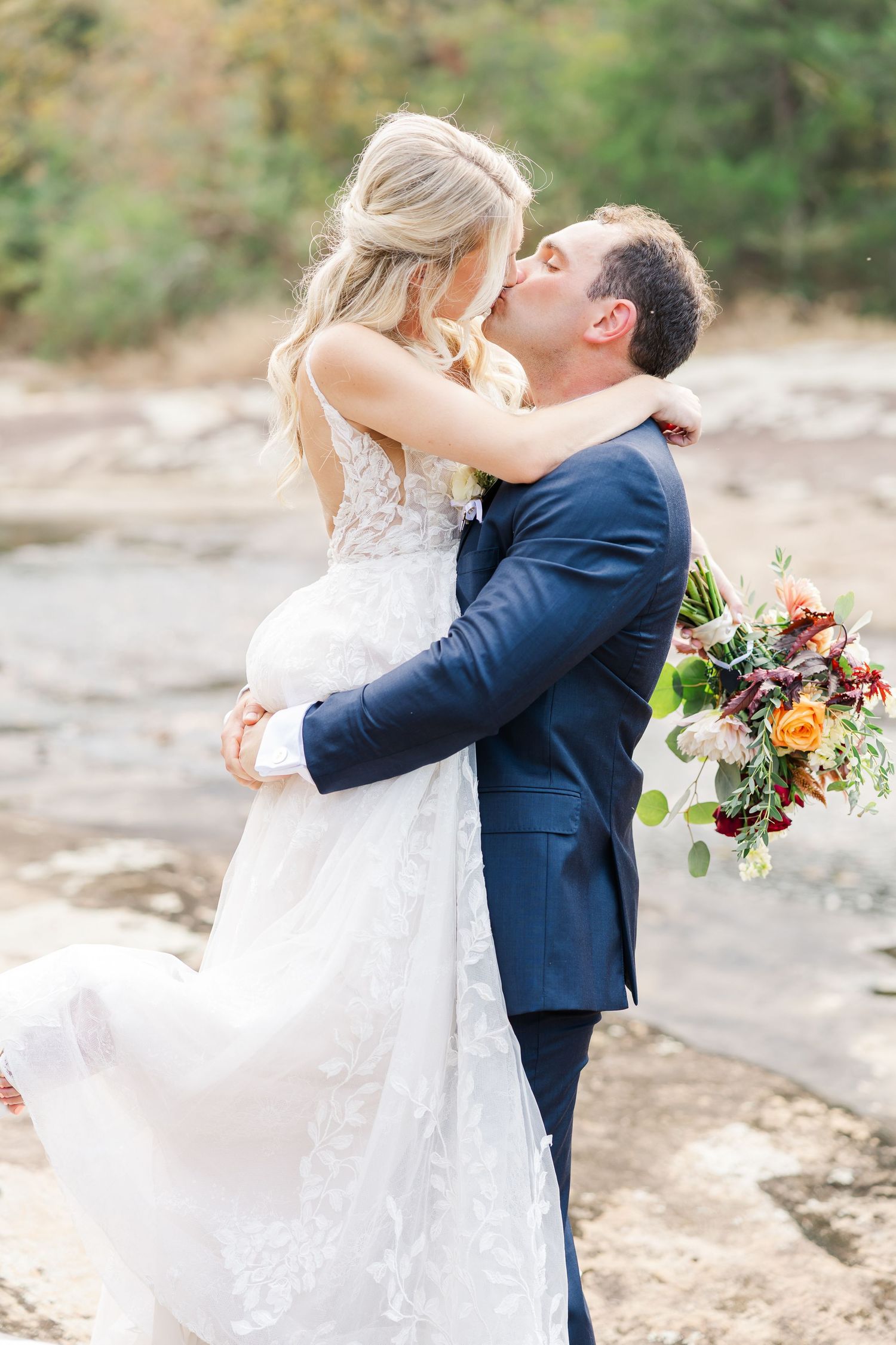 Romantic wedding couple shares a kiss with natural scenery by a creek and floral bouquet at The Mill at Fine Creek in Powhatan, VA