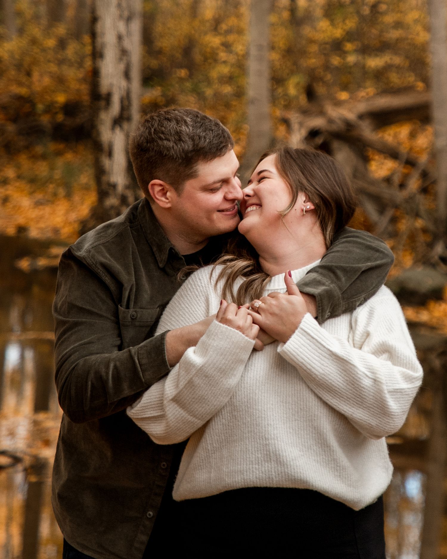 A couple shares a tender embrace and laugh together in an autumn forest setting with golden foliage in the background.