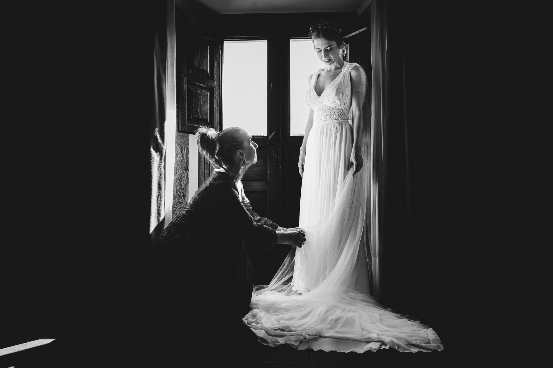 La novia ya vestida permanece de pie. Su madre ajusta el bajo del vestido antes de salir a la ceremonia en el Parador de Cangas de Onís.