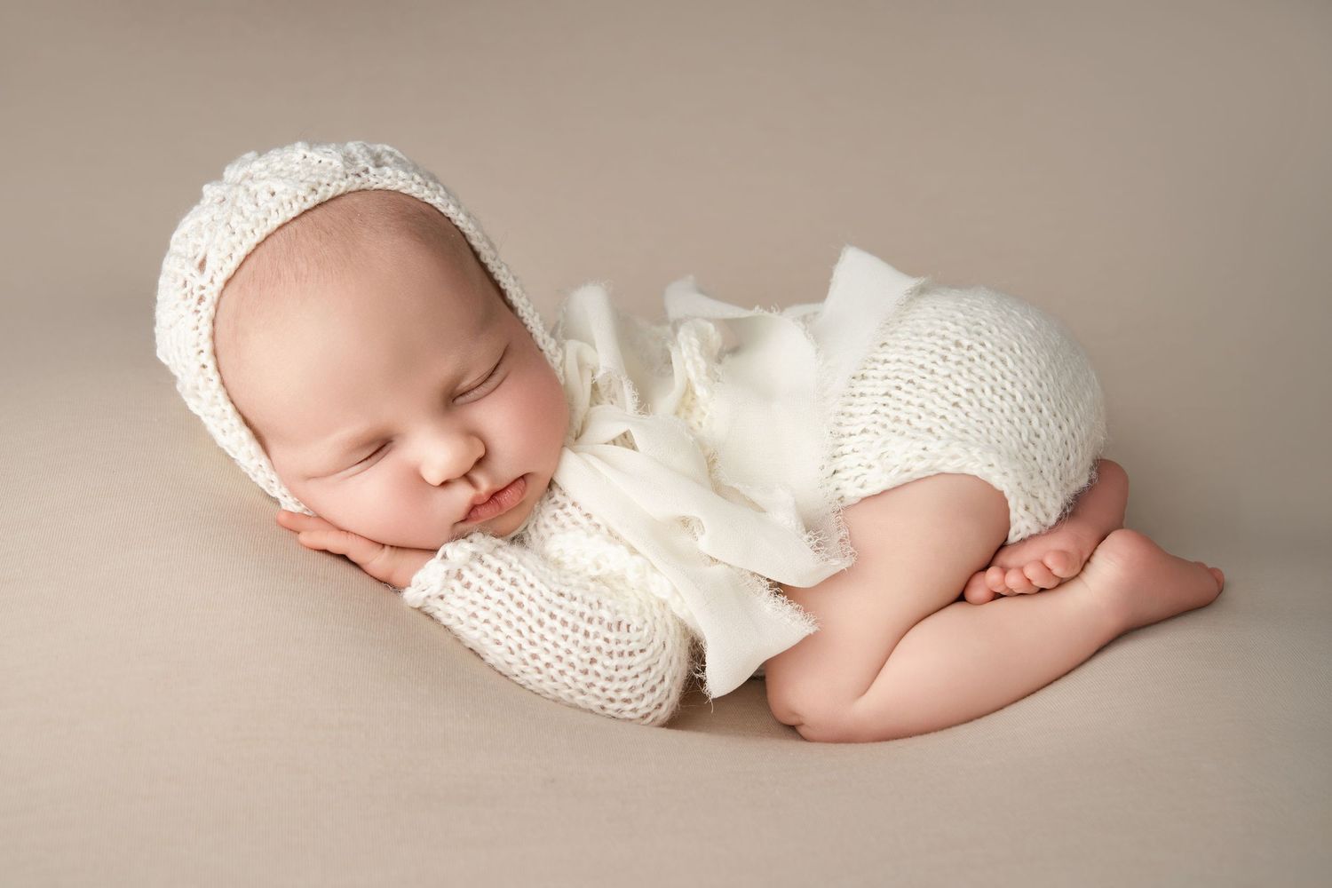 Peaceful sleeping newborn wrapped in cream colored knit on neutral backdrop.