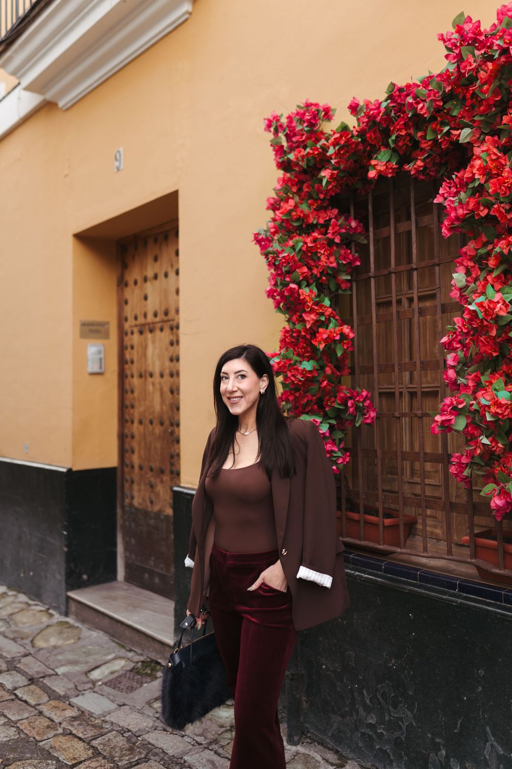 Woman in brown outfit stands by yellow wall with vibrant red bougainvillea flowers.