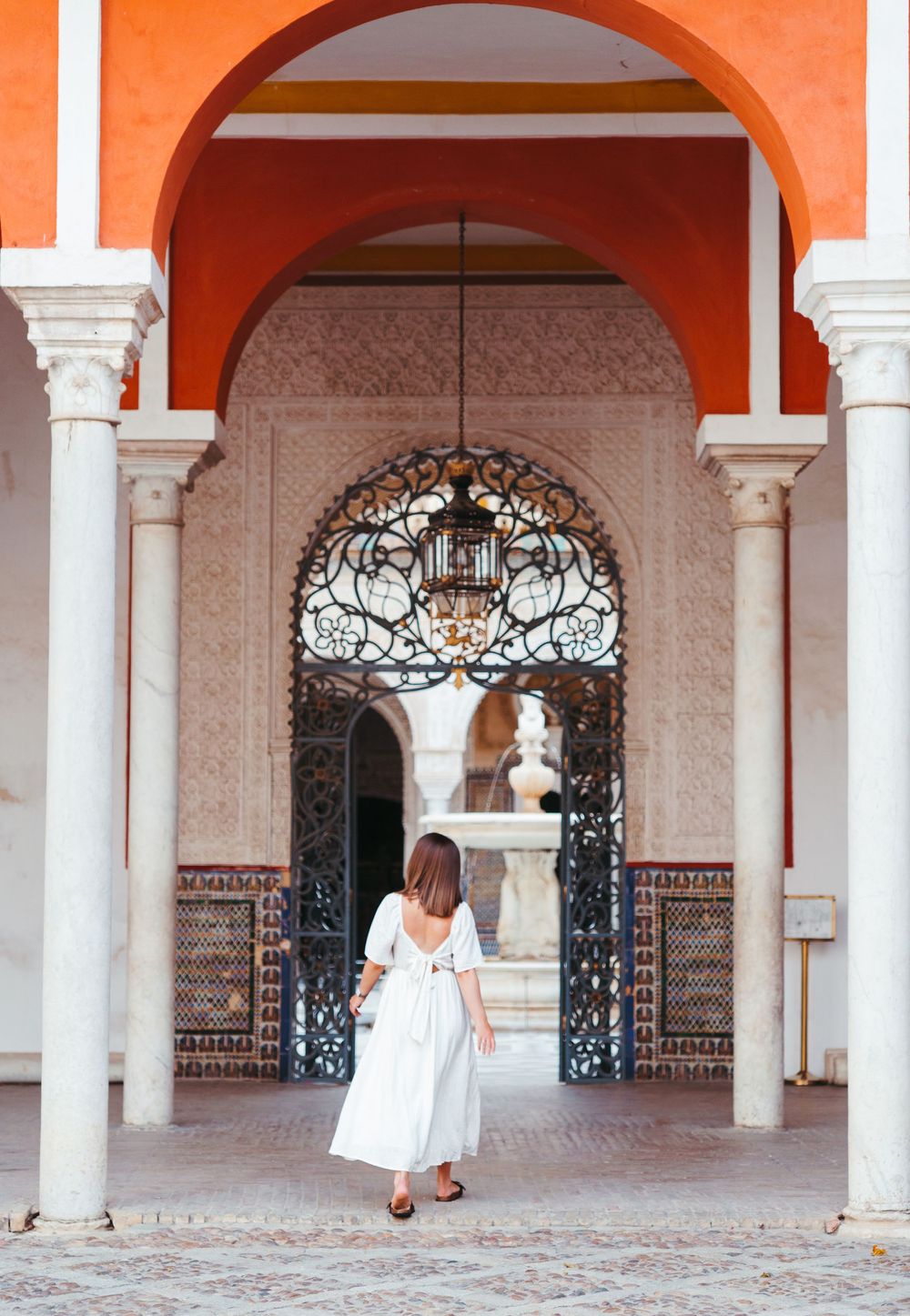Person in white dress walks through ornate orange archways with decorative tiles and wrought iron details.
