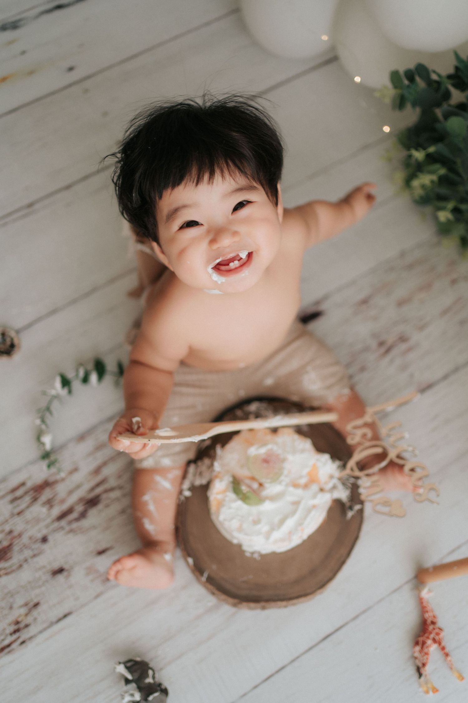 Baby in white smashes cake during first birthday photoshoot on rustic wooden floor.