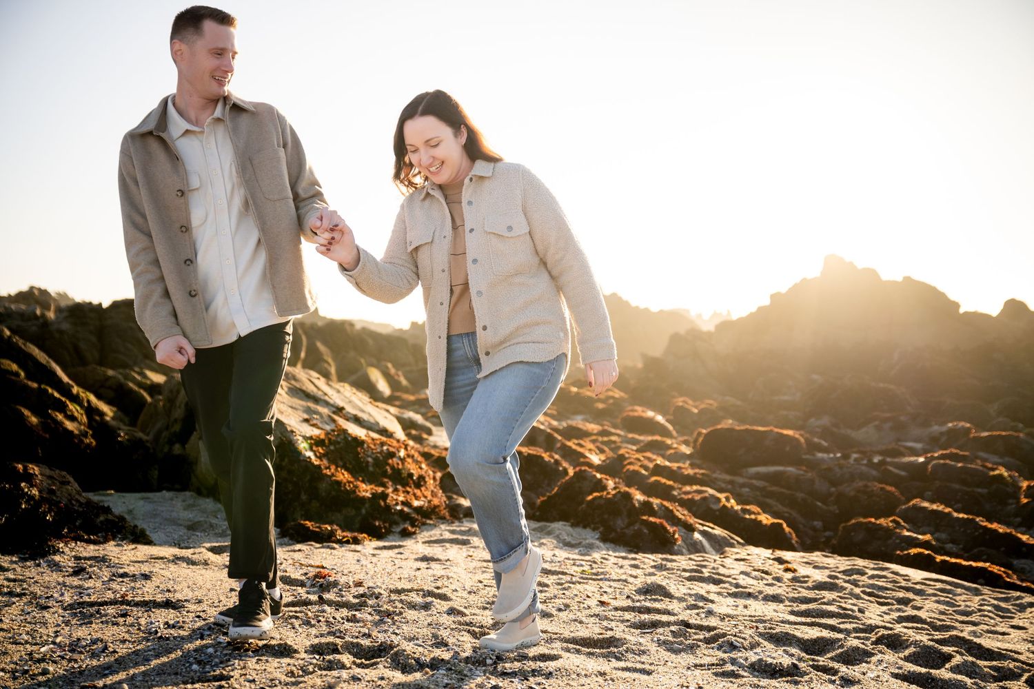 A couple walks hand in hand on a rocky beach at sunset, laughing and enjoying a carefree moment together.