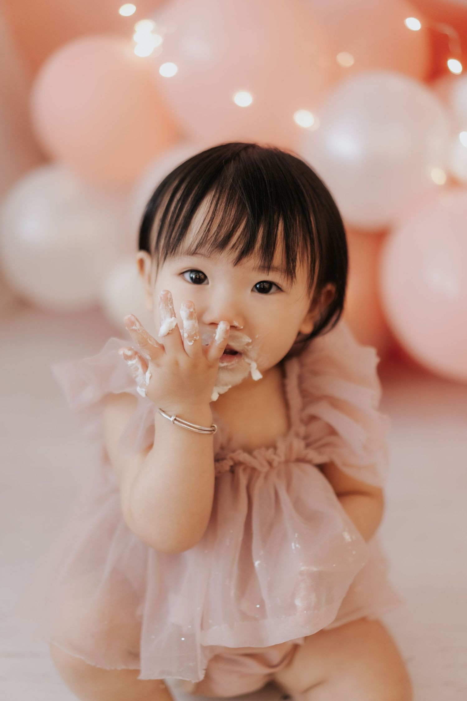Sweet baby in pink tulle dress sits among peach and white balloons with twinkling lights in background.