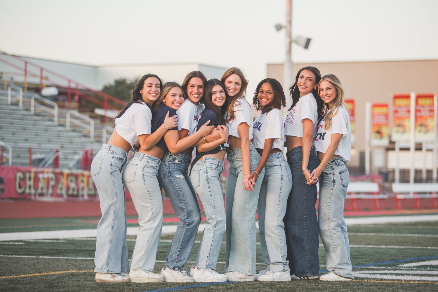 A group of friends wearing matching white t-shirts and blue jeans pose together on a football field at sunset.