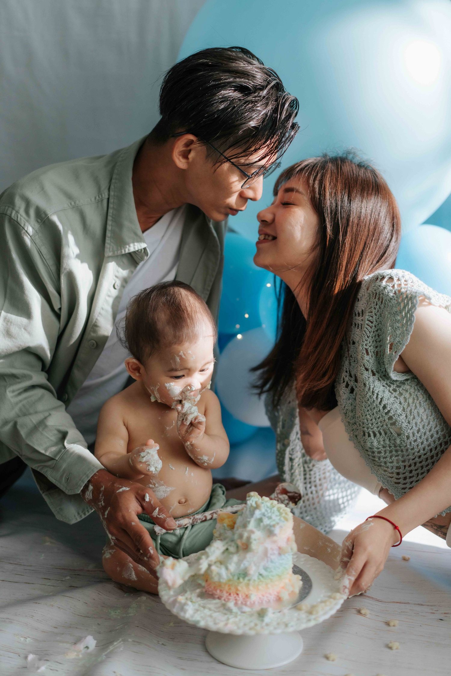 Family celebrates baby's first birthday with cake smash photo session in soft blue and white tones.