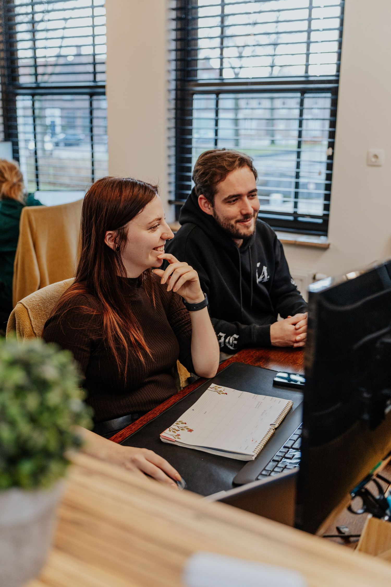 Two people collaborate at a desk while looking at laptop screens in a modern office setting with natural light.