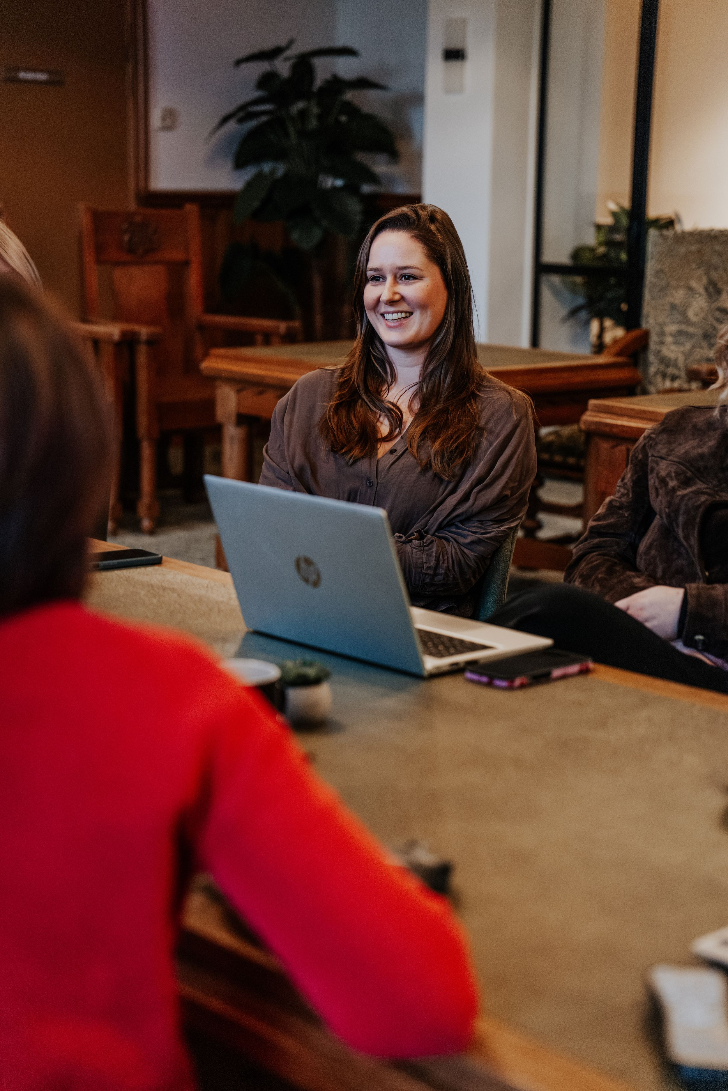 Someone works on a laptop at a wooden table in an office space with indoor plants visible in the background.