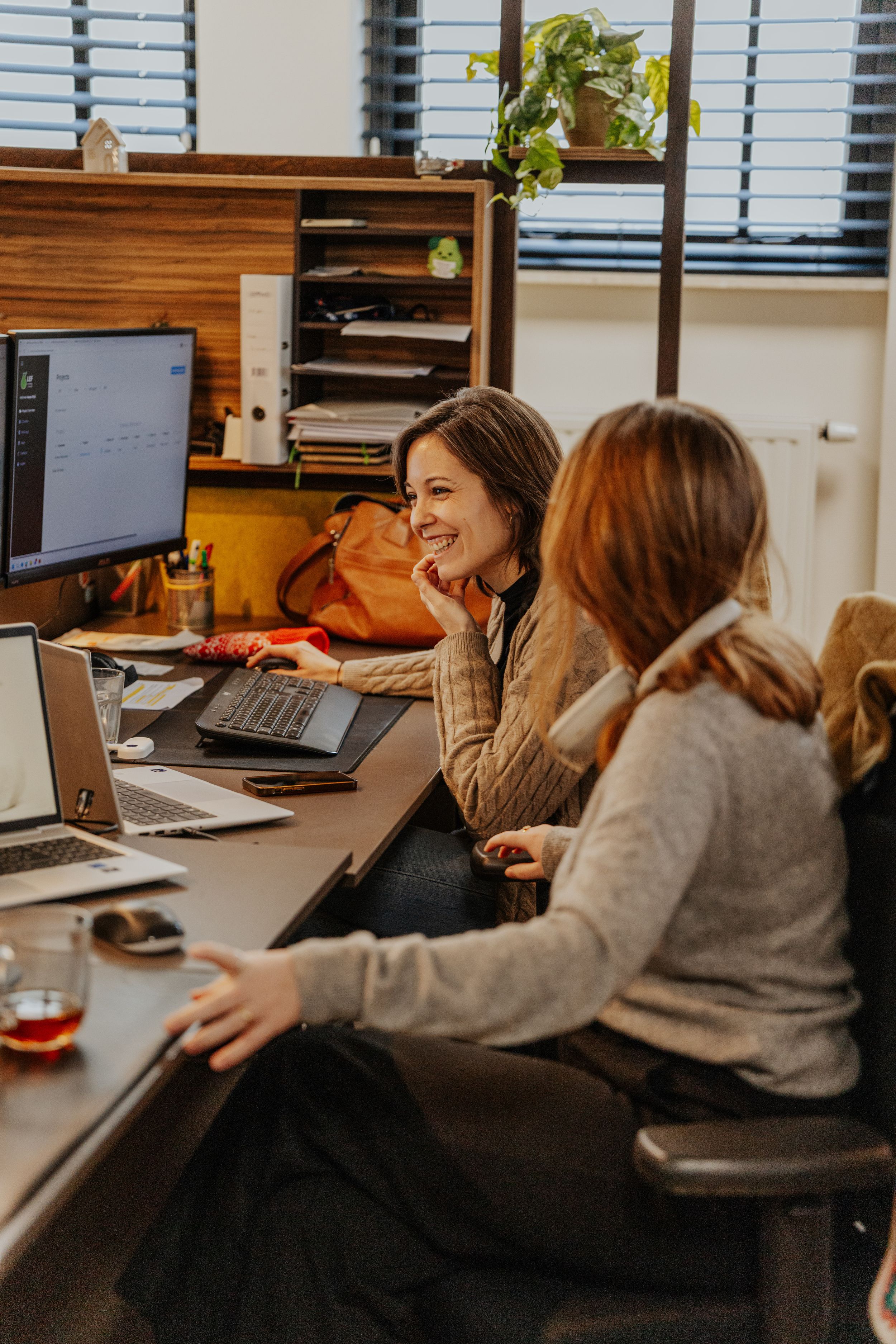 Two colleagues working together at a desk with computers in a cozy office setting with warm lighting.