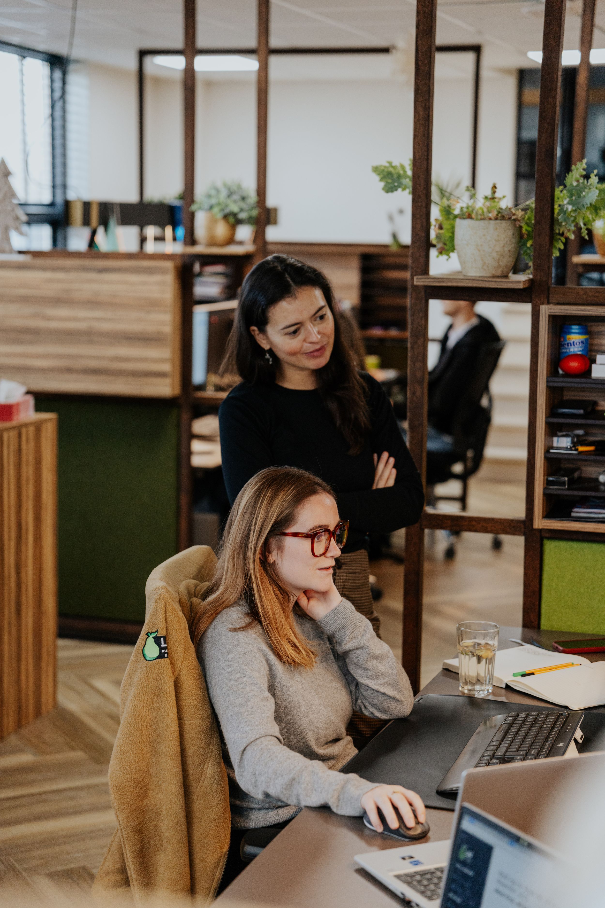 Two colleagues collaborate at a modern office workspace with wooden shelving and green accents.