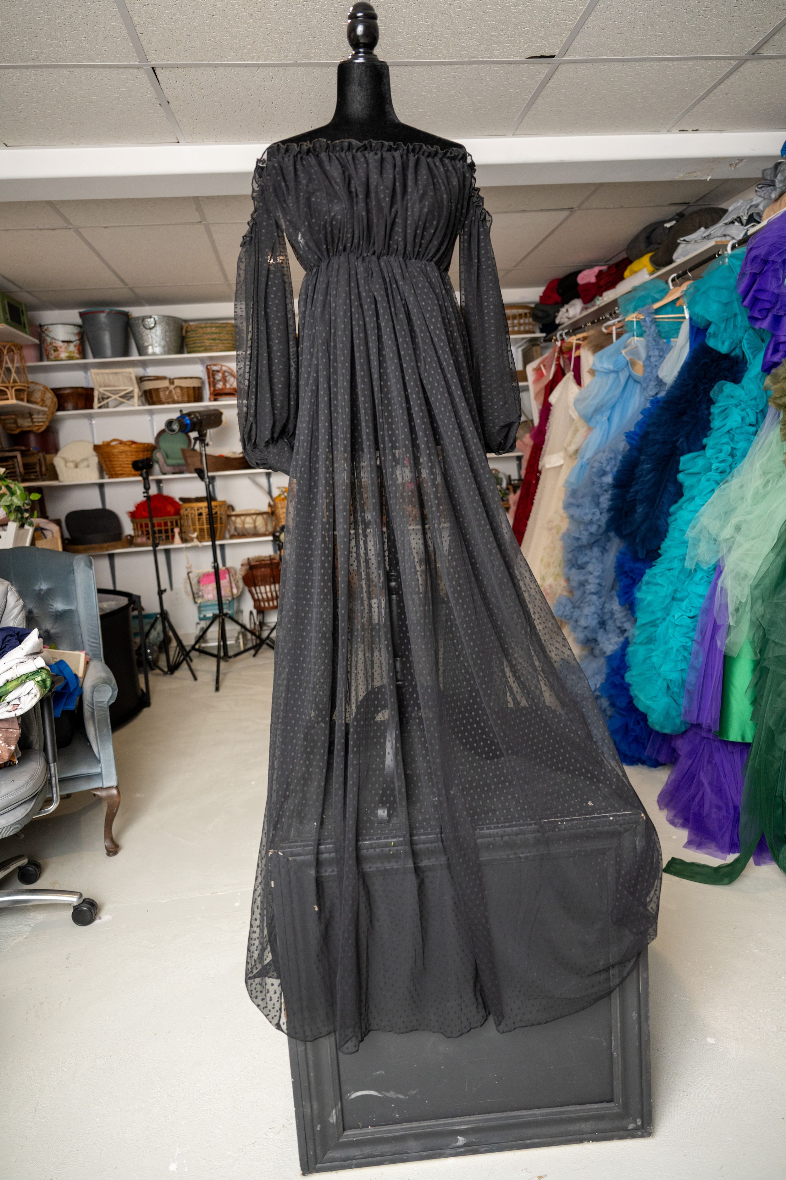 Black tulle dress with ruffled tiers displayed on mannequin in costume storage room with colorful garments visible.