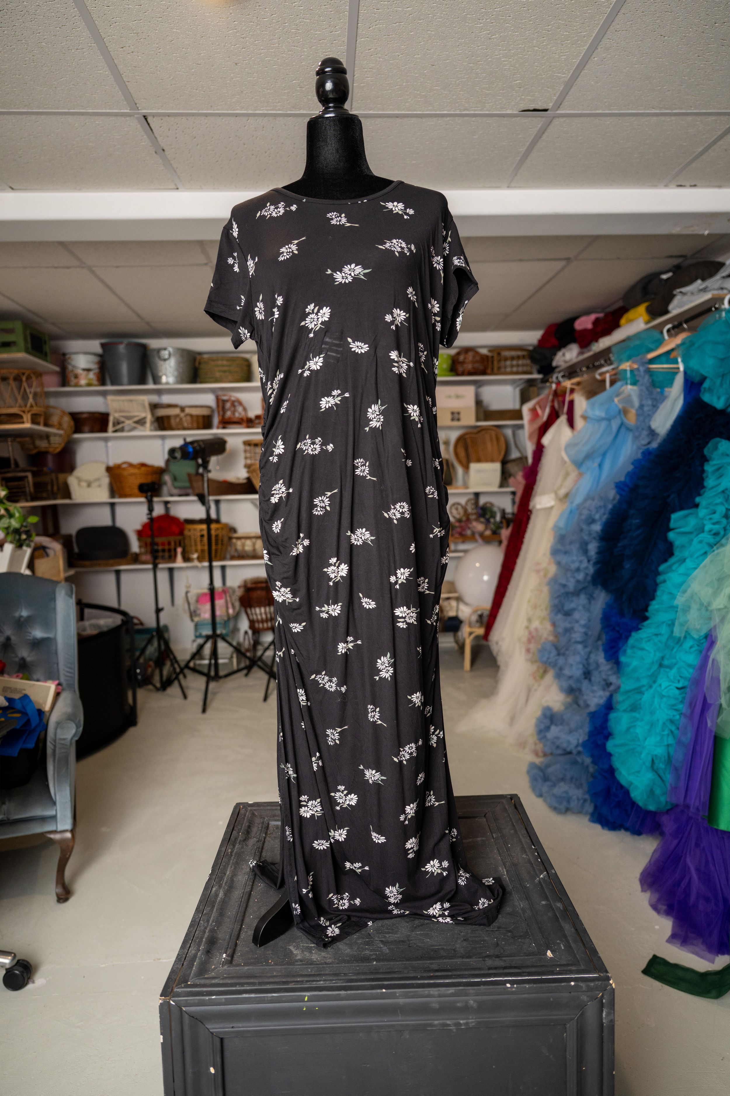 Black dress form displays a dark floral print dress in a costume storage room with colorful tutus visible in background.