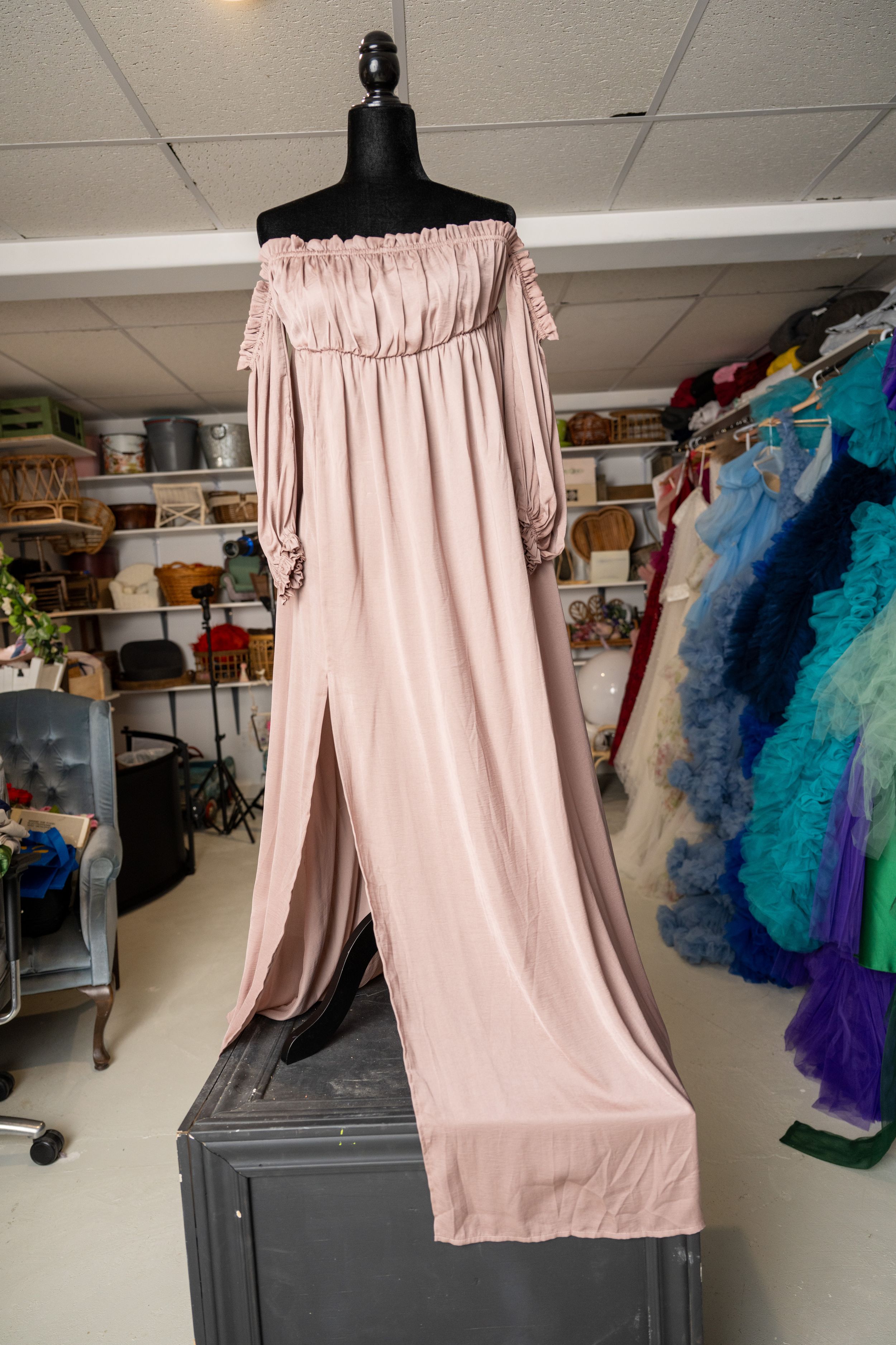 Pink off-shoulder dress displayed on a black mannequin in a studio workspace with colorful garments in background.