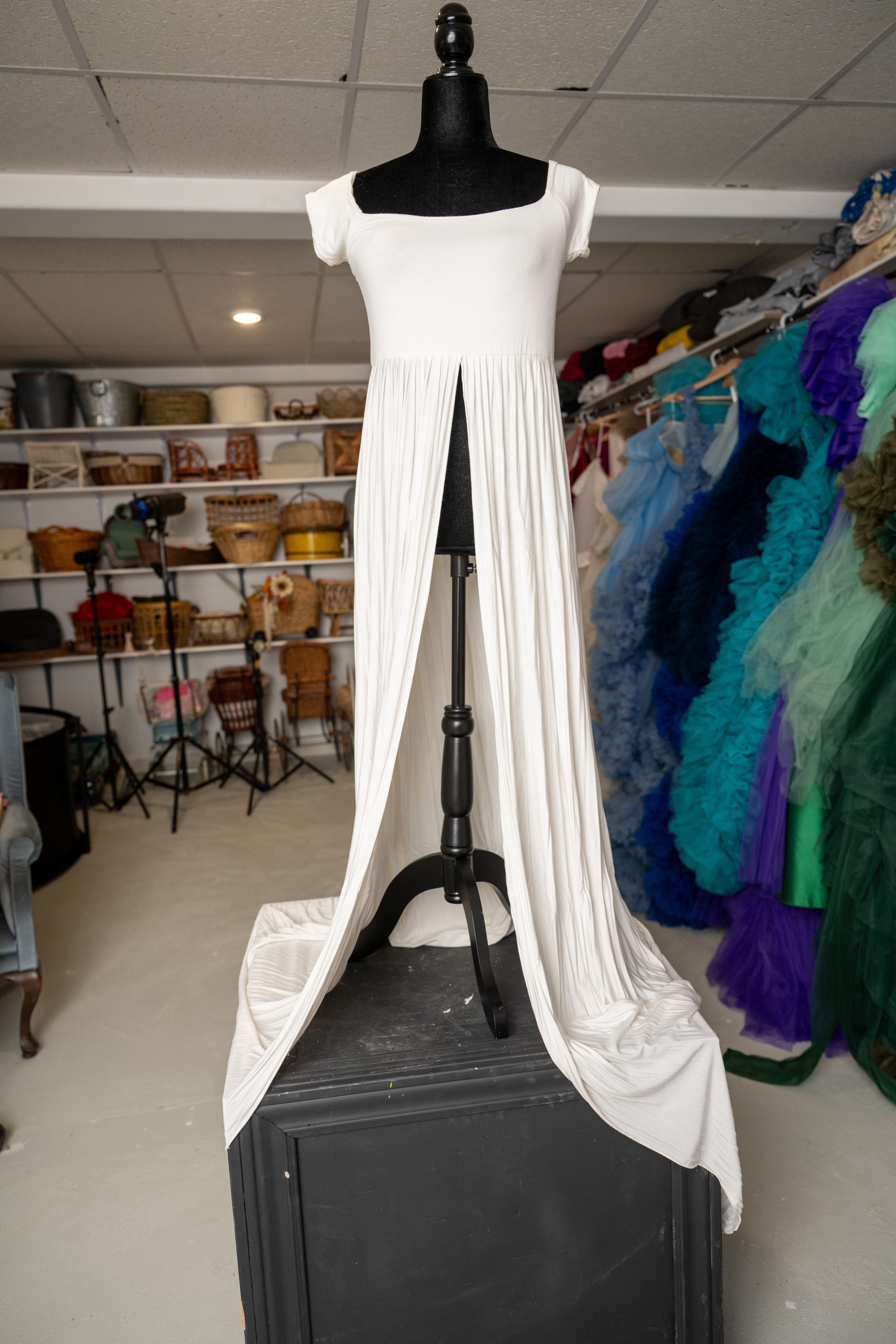 A white draped dress displayed on a black mannequin in a costume shop with colorful garments visible in the background.