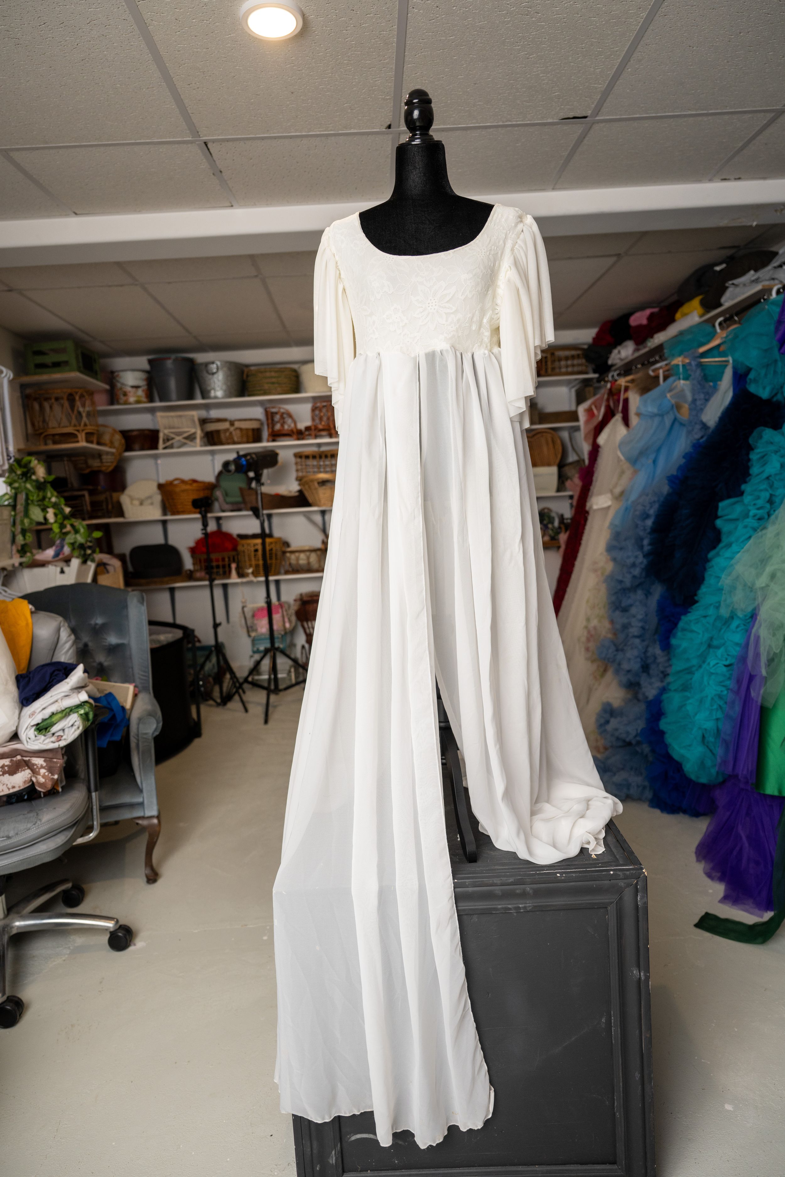 White flowing dress with bell sleeves displayed on a black mannequin in a costume storage room.
