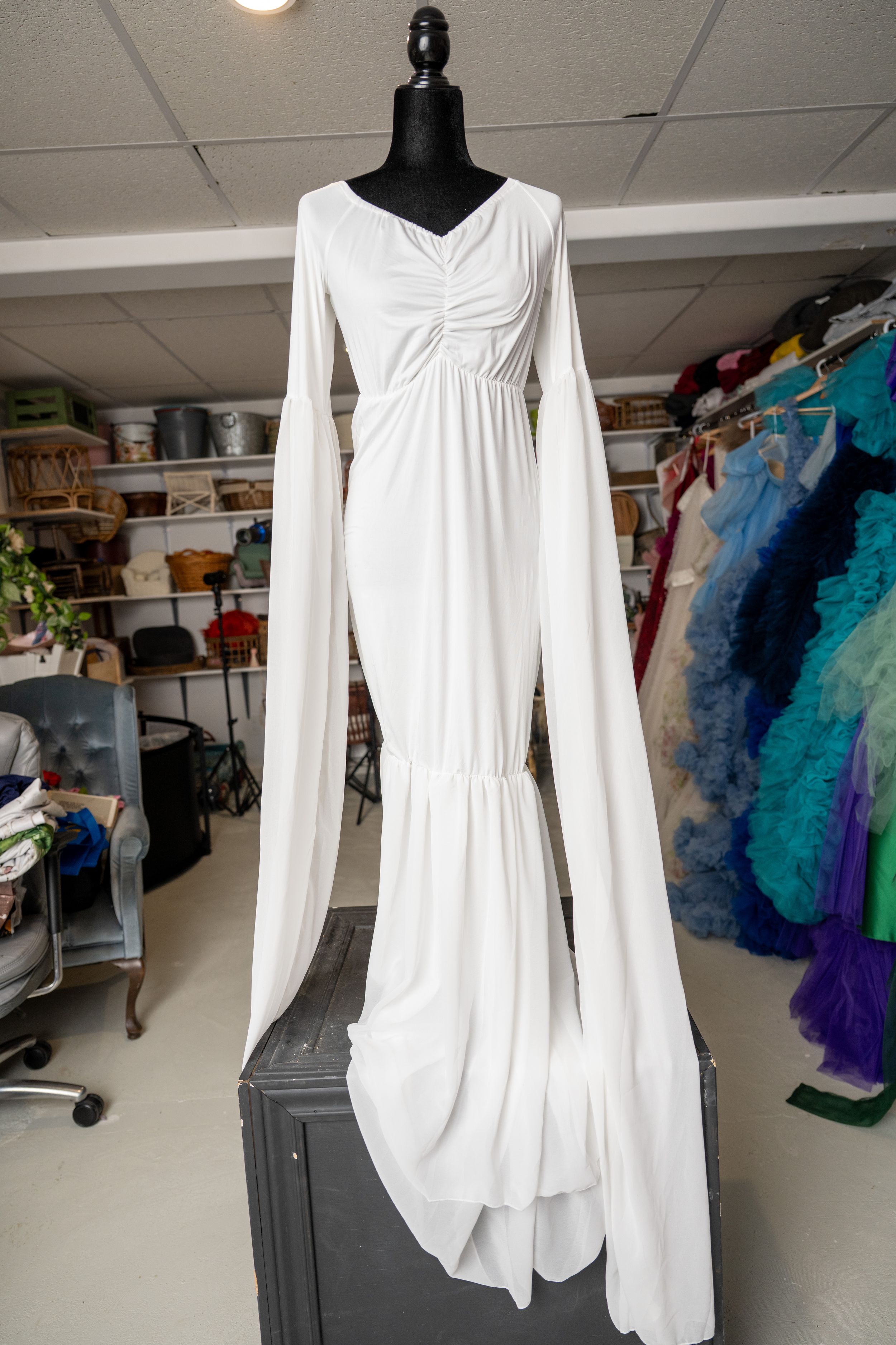 Long-sleeved white dress with gathered neckline shown on dress form against colorful backdrop.