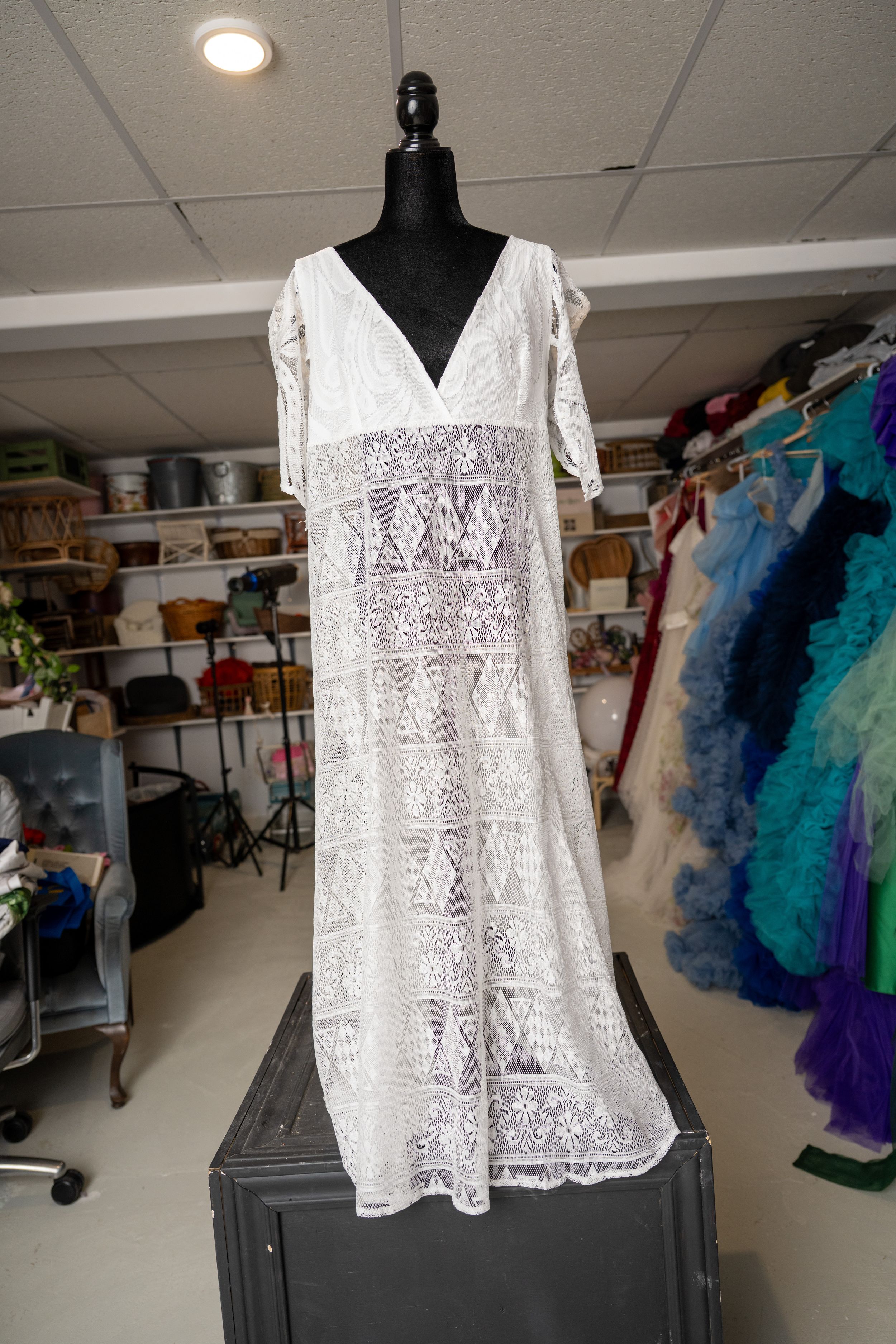 White lace peasant blouse displayed on a black mannequin form in a craft studio with colorful fabric in background.