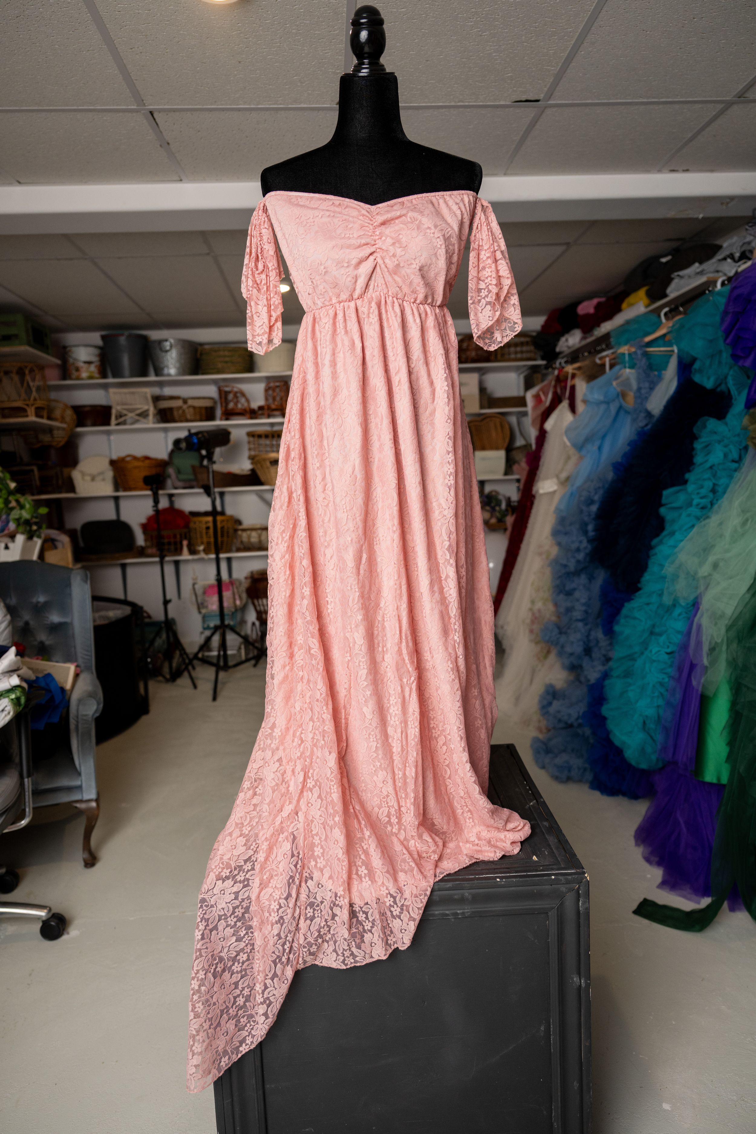 Pink off-shoulder lace dress displayed on black mannequin with colorful formal gowns visible in background.
