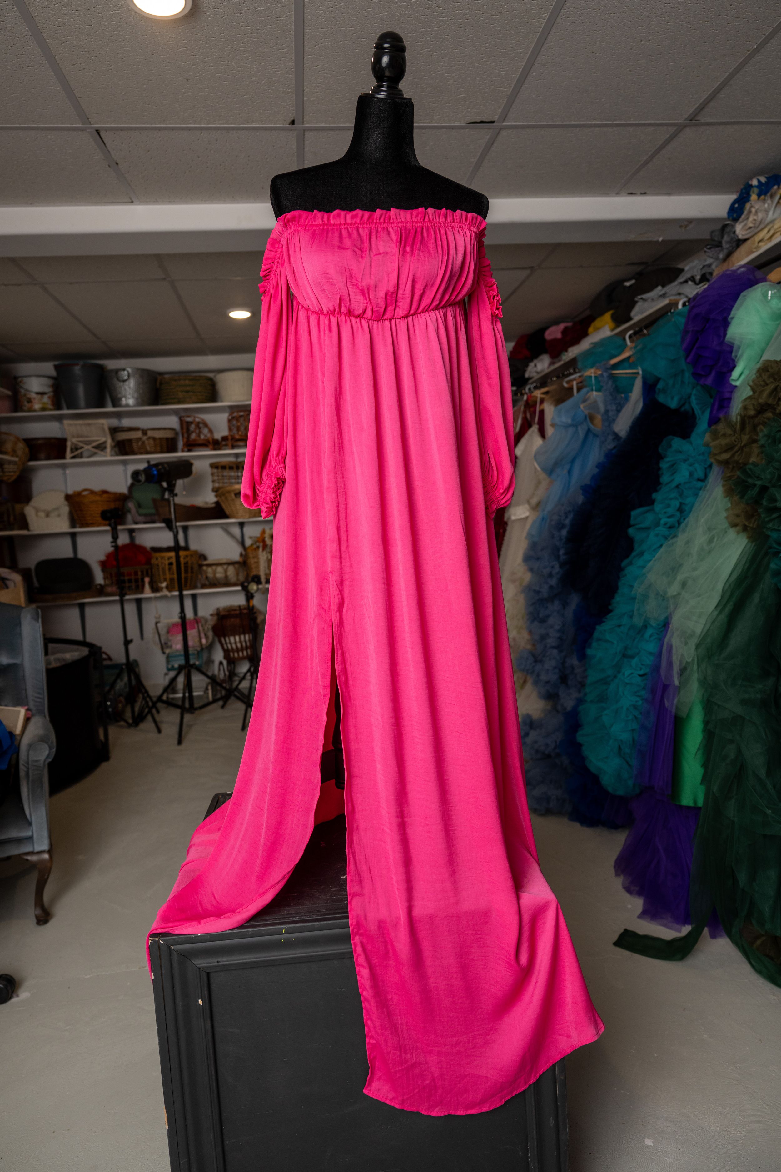 Hot pink off-shoulder formal gown with flowing fabric displayed on black mannequin in dress shop.