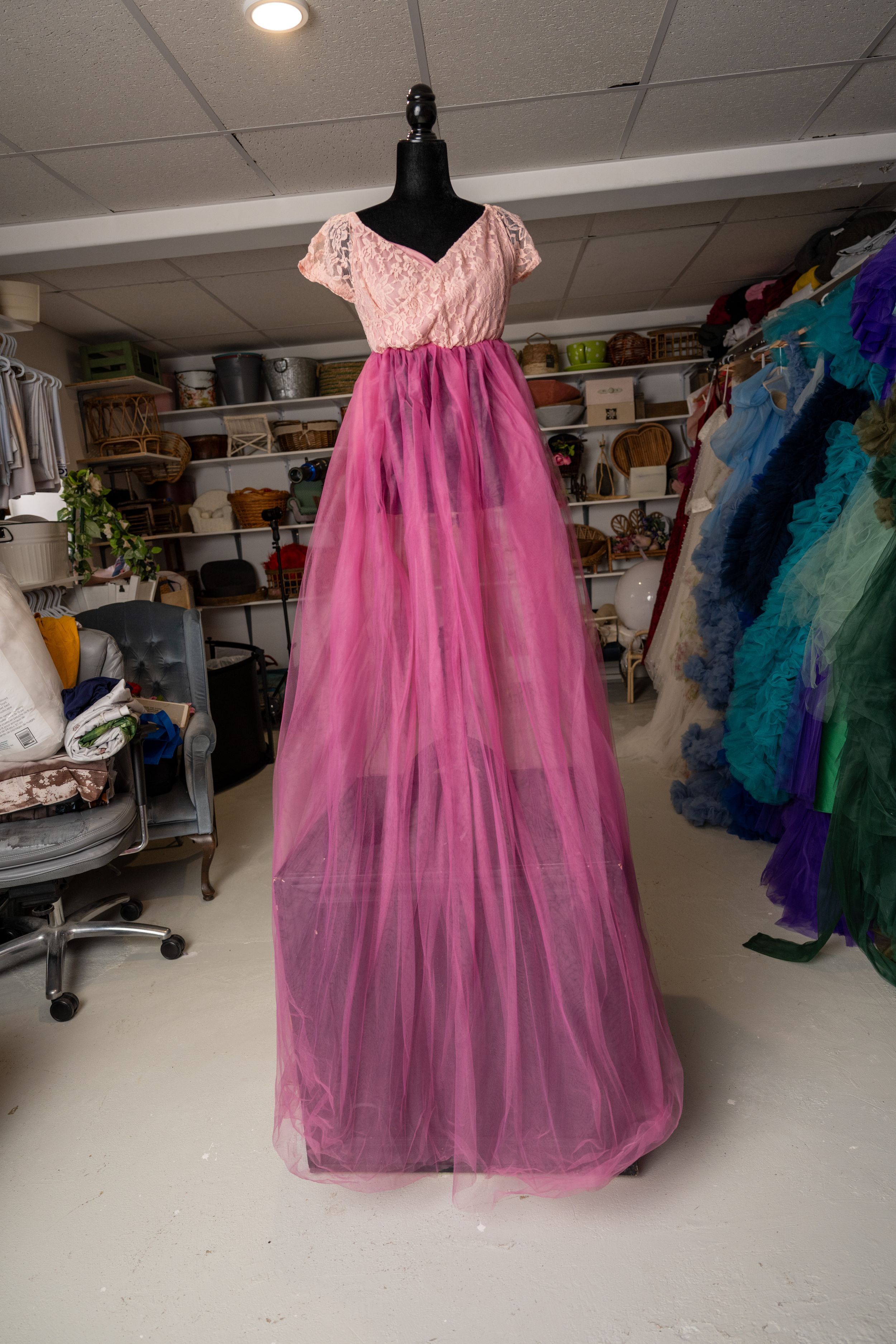 Pink tulle princess dress displayed on mannequin in costume shop with colorful garments visible in background.